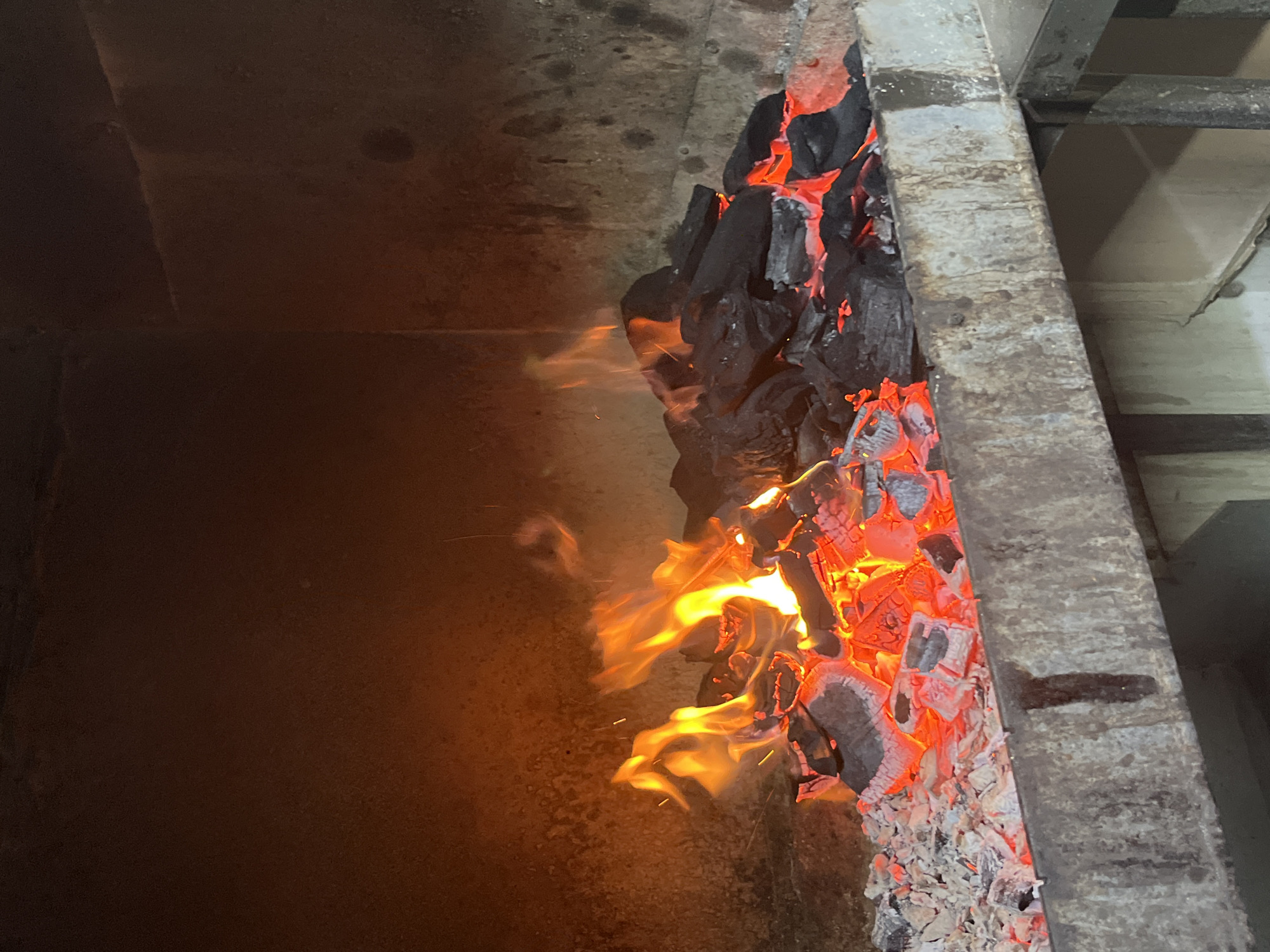 The image shows a close-up view of a charcoal grill in a dimly lit, enclosed space. The grill is filled with glowing red embers and black charcoal, with flames leaping from the burning fuel. The inside of the grill appears to be made of brick or stone, with a rough, textured surface. A metal grate or platform is visible at the bottom of the grill, holding the charcoal and embers. The overall impression is one of heat and intensity, with the flickering flames casting a warm glow on the surrounding surfaces. The scene is consistent with a barbecue or outdoor cooking area. There is no discernible location in the city of Dnipro, Ukraine.