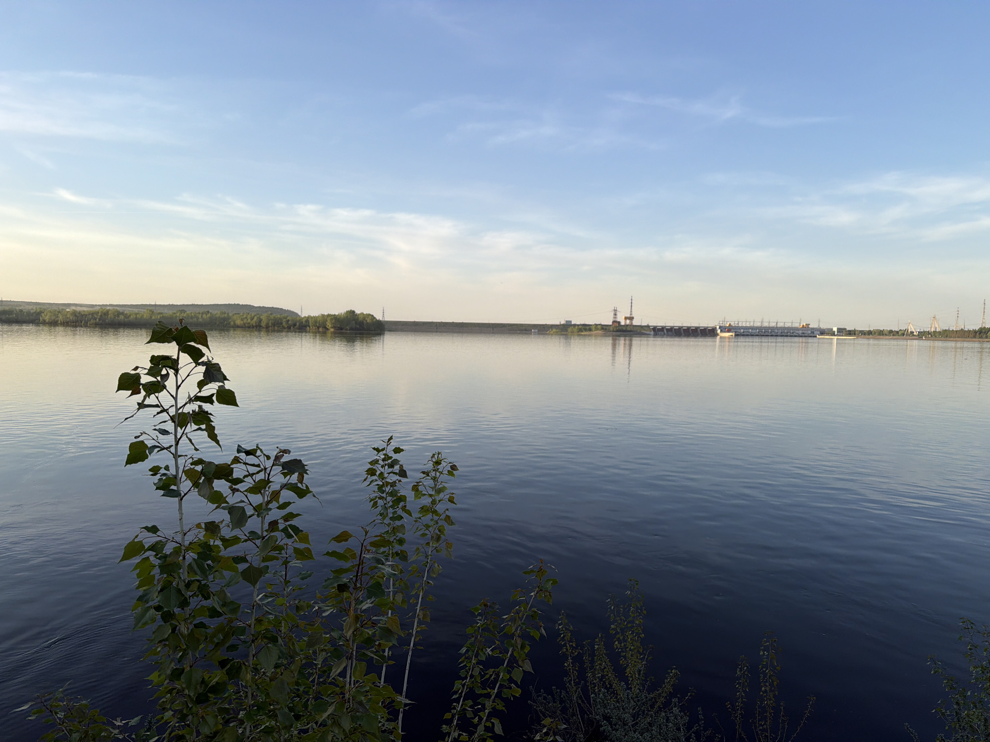 The image shows a wide river landscape under a light blue sky with wispy clouds. In the foreground, foliage partially obscures the view. The river's surface is mostly smooth, reflecting the sky. In the distance, a long dam stretches across the river, with structures and towers visible along its length. On the far bank, a line of trees is visible. The scene appears to be near the city of Noviy, Russia, based on the presence of the dam structure.