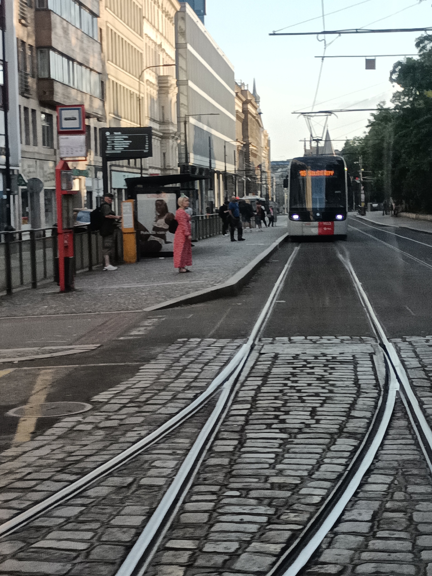 The image depicts a street scene in Prague, Czechia, likely a tram stop. In the foreground, tram tracks made of white metal are embedded in a cobblestone street. The tracks curve to the right, indicating a turn or junction. In the middle ground, a modern tram is approaching, its headlights on. The tram displays "15 Smíchov" on its front, indicating its route. On the left side of the tram tracks, a raised sidewalk features a bus stop with a sign and a red pole. Several people are visible on the sidewalk, including a woman in a long pink dress and a man standing near the bus stop. Behind the sidewalk, buildings with modern architectural elements and older facades line the street. The environment suggests an urban setting with active public transportation.