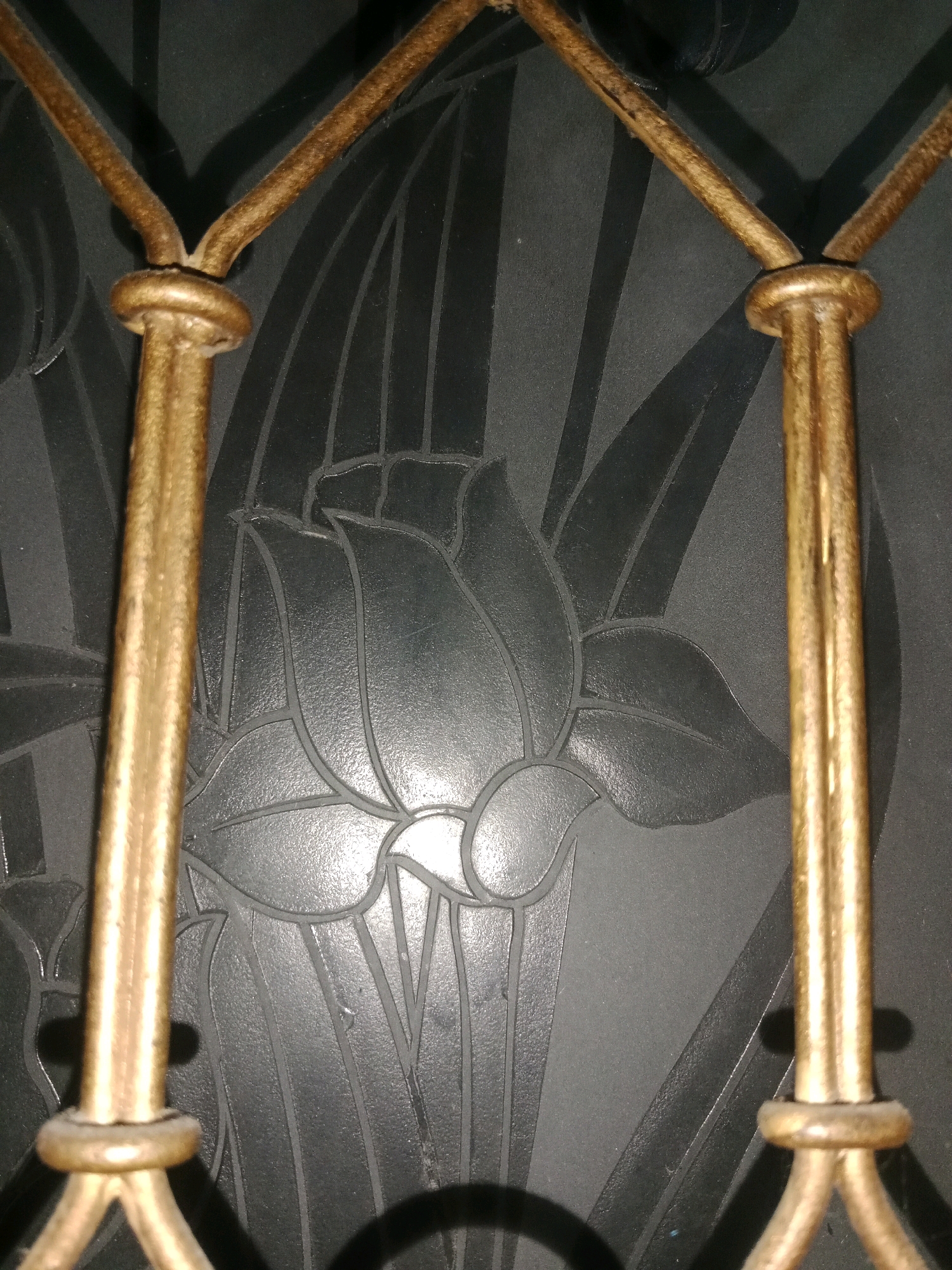 A close-up, low-angle, indoor shot captures a gold metal decorative element against a dark background featuring an embossed floral pattern. The decorative element consists of two vertical posts, each connected by a decorative knot near the top, and then extending upwards and outwards into a Y-shape. The floral pattern appears to be a stylized lotus flower with broad leaves. Shadows are cast on the background by the metalwork. The image's location is identified as Ringinrejo, Indonesia, though no contextual clues within the image itself support this specific location.