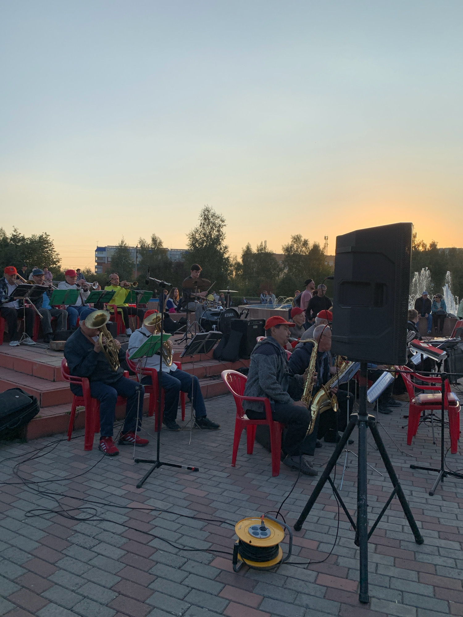 This photograph captures a performance by an orchestra outdoors, likely in Prokopyevsk, Russia, during the golden hour as indicated by the warm sky. The orchestra members, predominantly older men wearing red caps, are seated on tiered red steps and red plastic chairs, playing various brass and wind instruments including tubas, saxophones, and trumpets. Some members have music stands in front of them. In the background, trees, a building, and a fountain can be observed, suggesting a park or public square setting. Several people are seated or standing in the background, observing the performance. A large black speaker is prominently positioned in the foreground, next to a tripod stand. Electrical cables are visible on the paved ground, connected to a yellow cord reel. The overall scene depicts a community event or concert.