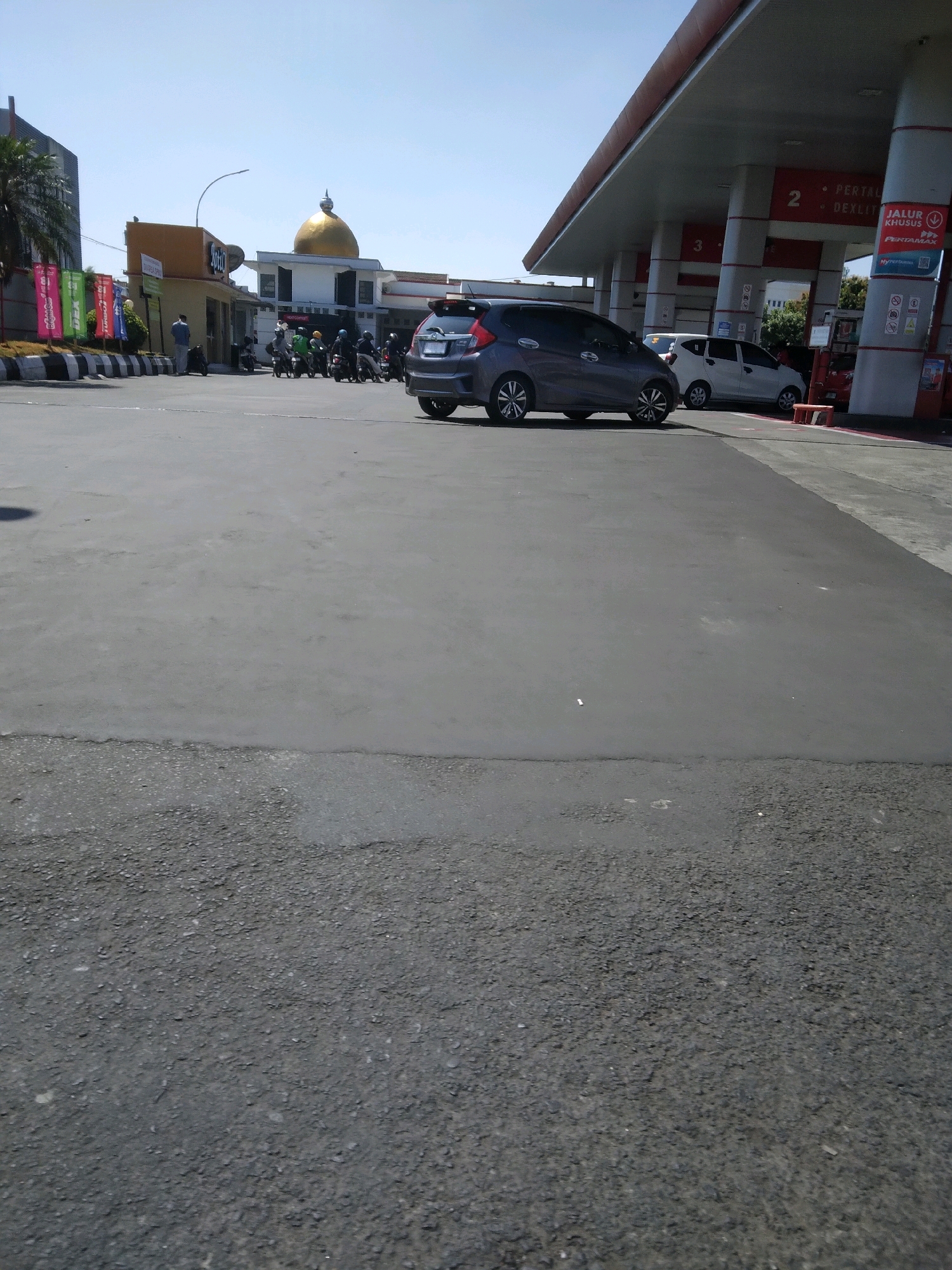 A wide-angle, eye-level outdoor shot captures a Pertamina gas station in Purwokerto, Indonesia, on a clear, sunny day. The foreground is dominated by a gray asphalt road, transitioning to a smooth concrete surface at the midground. In the midground, a dark gray hatchback, possibly a Honda Jazz, is parked under the gas station's canopy. To the right of the hatchback, a white sedan is also visible beneath the canopy. The gas station features red and white pillars supporting a large, angled roof. Several gas pumps are lined up, with signs indicating fuel types and lanes. Behind the gas station, a building with a distinctive golden dome, likely a mosque or a prominent landmark, is visible. A row of motorbikes is parked in front of a yellow building to the left of the dome, with several people standing nearby. Colorful banners are displayed on poles in the far left of the scene. The sky is clear blue.