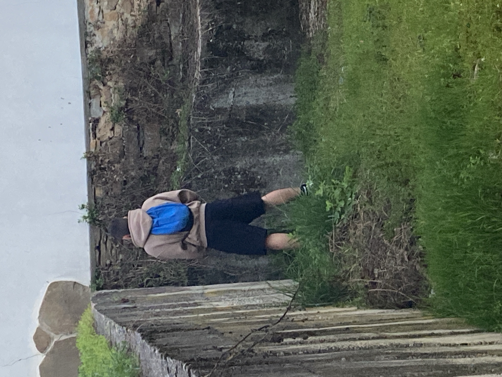 A person wearing a tan hoodie, black shorts, and sneakers with a blue backpack is lying on the ground next to a stone wall and overgrown grass. The location is likely near a stone structure in Troyan, Bulgaria, given the prompt. The scene consists of natural elements like grass, stone, and a person resting in this environment.