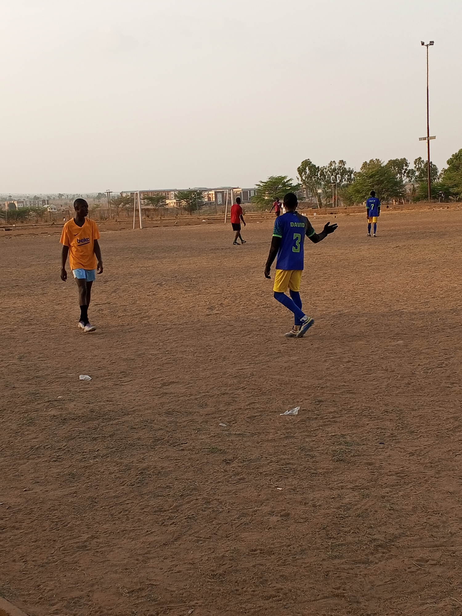 The image shows a group of people playing soccer on a dirt field in Minna, Nigeria. A person in an orange jersey with the word "Beko" is walking on the left side. In the center, another person wearing a blue and yellow jersey with the name "David" and number 3 on the back is walking and gesturing with their left hand. A third person wearing a red jersey is walking in the background, and a fourth person wearing a blue and yellow jersey with the number 7 on the back is standing on the right. There are soccer goals in the background and a tall light pole on the right. There is a building in the distance.