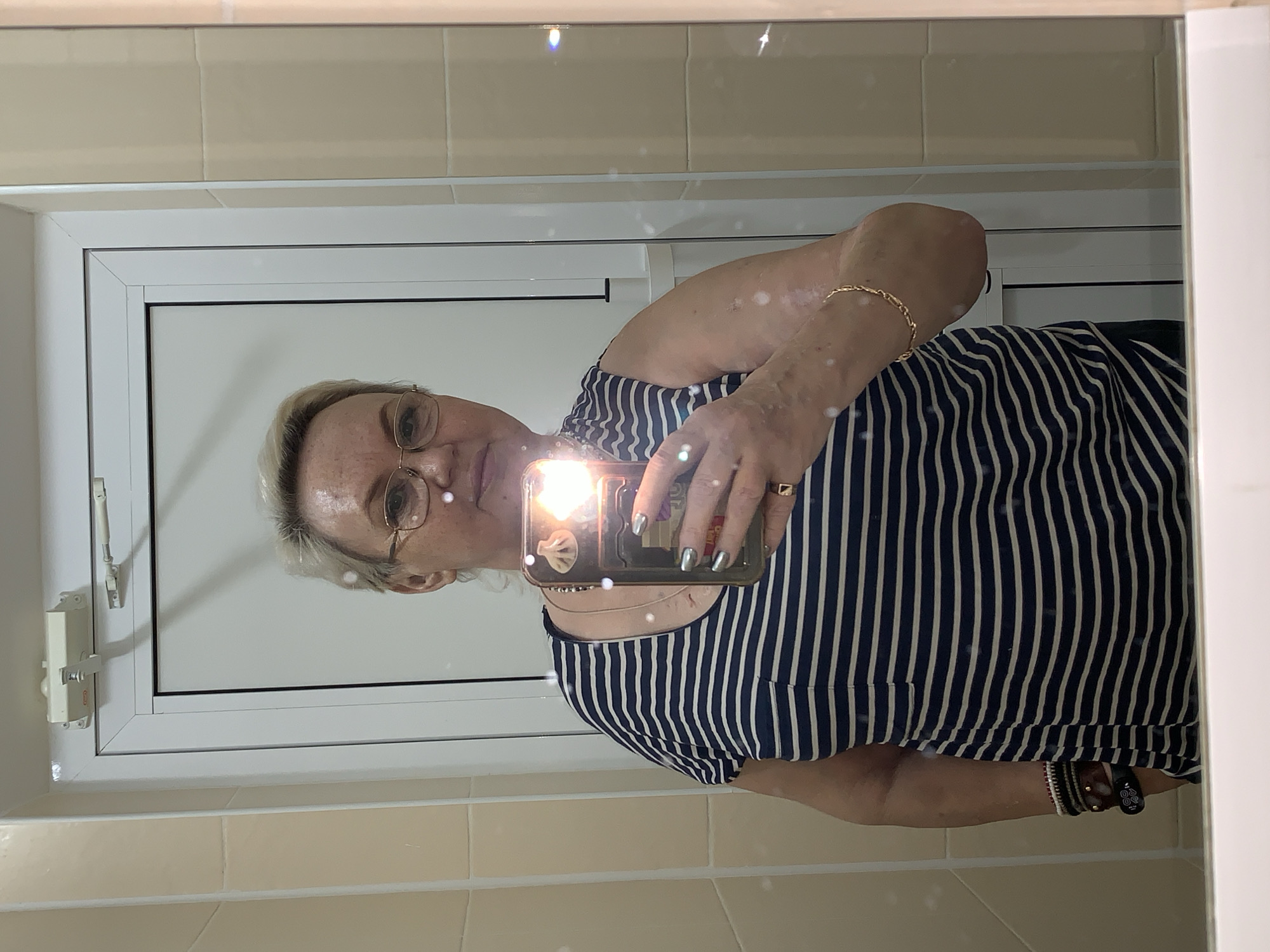 A mirror selfie taken indoors in Chaikovsky, Russia, features a person with light-colored (blonde or gray) hair and glasses. The individual is wearing a dark blue and white striped sleeveless top. They are holding a smartphone, which is reflected prominently in the mirror, with its flash creating a bright glare that partially obscures the face.
The person's left wrist sports a gold chain bracelet, while their right ring finger wears a gold-toned ring. On their right wrist, multiple bracelets are visible, including what appears to be a fitness tracker or smartwatch displaying faint numbers, possibly "40" or "80." The smartphone is encased in a protective cover that shows a small, light-colored icon, possibly a seashell.
The background visible in the reflection consists of light beige or cream-colored tiles, indicating an indoor setting, likely a bathroom or hallway. A white window or door frame with a latch mechanism is also reflected behind the person. The overall lighting appears to be artificial, given the prominent phone flash. No strong emotions are discernible from the person's expression due to the camera's angle and flash.