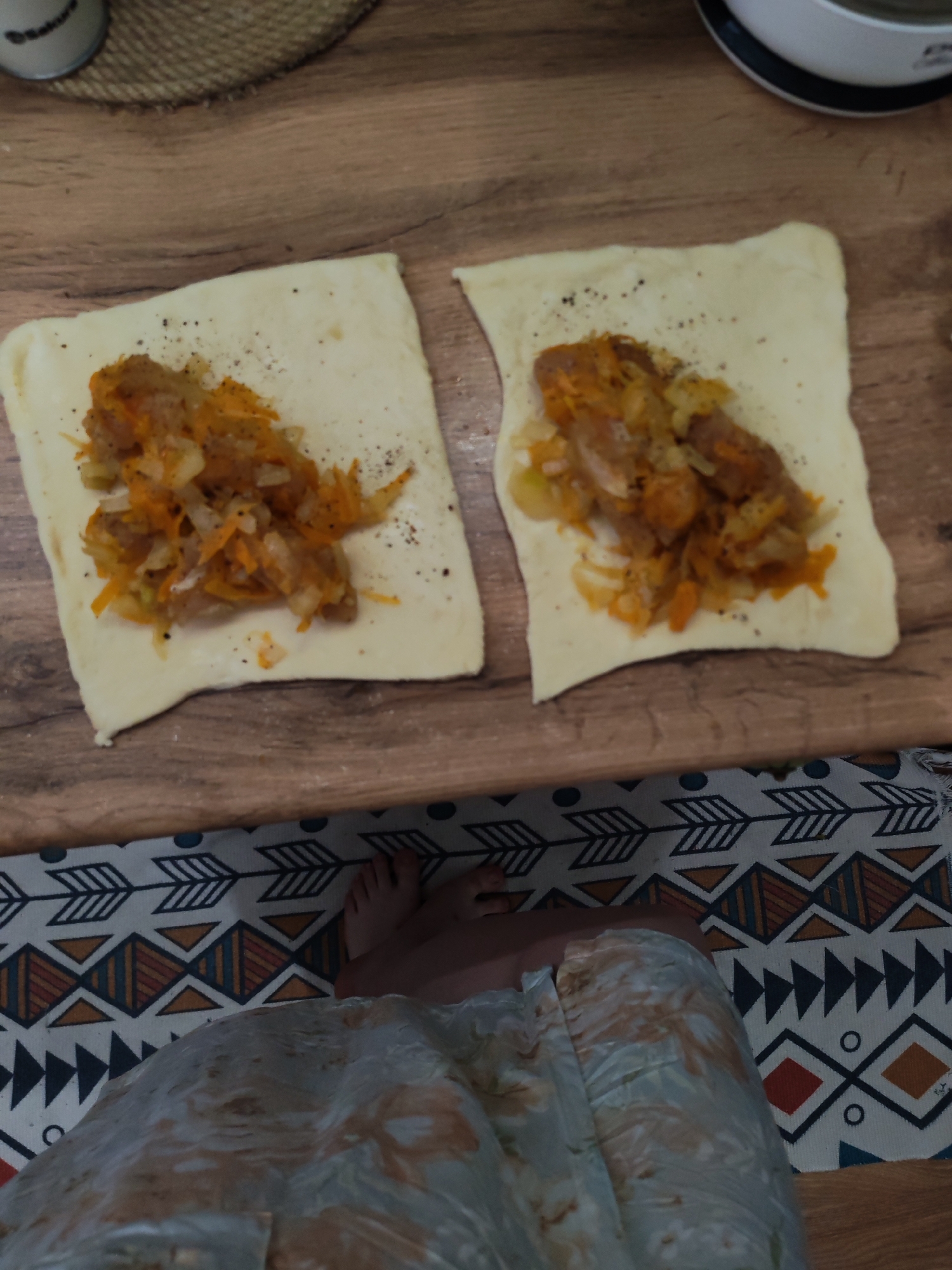 The image captures a scene of food preparation on a wooden surface, likely a kitchen counter or cutting board. Two rectangular pieces of raw dough are laid out, each containing a savory filling. The filling appears to be a mixture of cooked vegetables, prominently featuring shredded carrots and diced onions, combined with pieces of meat or sausage. Black specks, likely ground pepper, are sprinkled over the filling and dough.
In the upper left corner, a portion of a dark container with a light-colored lid, possibly a spice shaker, is visible on a woven mat. Partially visible text on the container reads "Satur...". In the upper right, a white object with a black rim is partially seen, possibly a part of a kitchen appliance or dish.
At the bottom of the frame, a person's bare feet and lower legs are visible, resting on a patterned rug. The rug displays a geometric design with arrows, triangles, and diamonds in white, black, orange, and blue. The person is wearing light-colored shorts or a skirt with a mottled pattern in hues of light blue, white, and orange-brown. The scene suggests a home setting in Izhevsk, Russia.