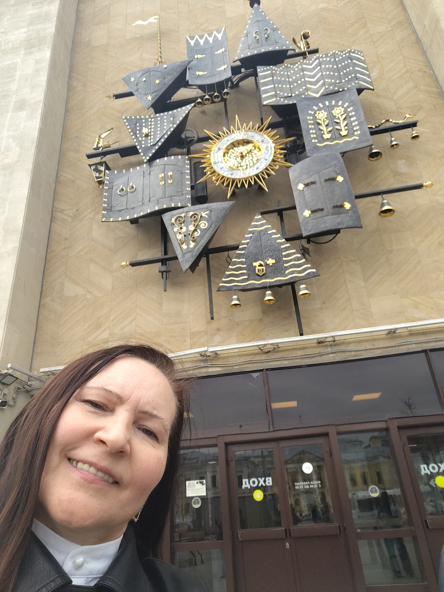 The image shows a woman posing for a selfie in front of a building with a large decorative clock on its facade. The woman is wearing a black leather jacket over a white collared shirt. She has long brown hair and is smiling.
Behind her, a modern building has a facade covered with a light brown or beige stone paneling. The focal point is a large, complex clock made up of various geometric shapes and decorative elements, including bells. The clock’s design incorporates triangles, squares, and what appears to be a central sun-like figure.
The building has a set of glass doors with the word "ДОХА" written on them. The reflections in the glass doors show the interior of the building and the activity inside. Wires and conduit run along the wall near the doors.
The location is in the city of Unknown, Unknown, as the signs on the building are written in Cyrillic.