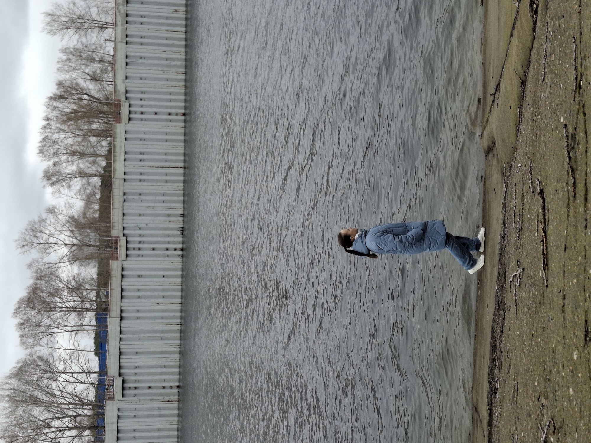 The image shows a woman standing on the shore of a body of water, likely a river or lake. The location is likely in the city of Chaikovsky, Russia. The woman is wearing a blue jacket, jeans, and white sneakers. She has dark hair pulled back in a ponytail. To the left of the frame is a concrete wall with bare trees behind it. The sky is overcast. The water appears calm, with slight ripples on the surface. The shoreline is a mix of sand and concrete. Some roots are coming out of the concrete.