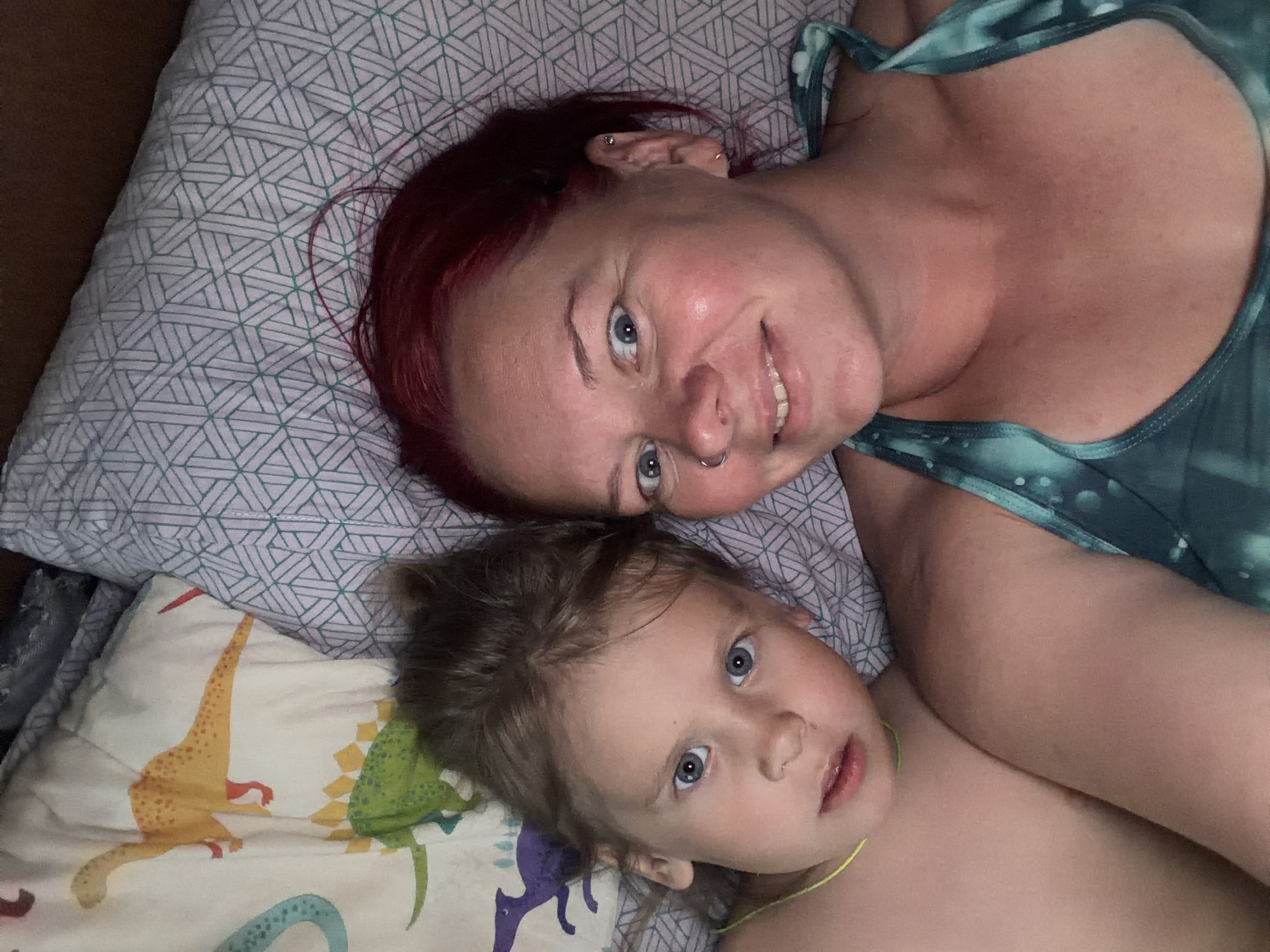 An adult female and a young child are depicted lying in a bed within a residence located in the city of Chaikovsky, Russia. The adult female is positioned on the right, presenting a front-facing view, with red/auburn hair and a light-colored complexion. She exhibits a nose ring in her left nostril and multiple piercings on her left ear. Her eyes are open and directed towards the camera, and she displays a slight smile. She is wearing a teal-green patterned tank top or sports bra. To her left, a child, approximately 3-7 years old, also faces forward. The child has light brown, tousled hair and blue eyes, with a neutral expression. A thin, neon yellow-green cord or necklace is visible around the child's neck. The child appears to be shirtless. Both individuals are lying on pillows; the pillow behind the adult features a geometric pattern of interlocking lines, while the pillow behind the child displays cartoon dinosaur illustrations, including an orange long-necked dinosaur, a green spiked dinosaur, and a purple winged dinosaur. The lighting is ambient.