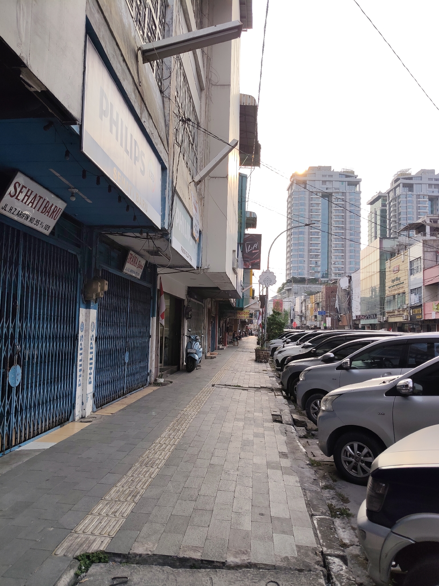 This is a street-level, eye-level, long shot of a street in the city of Medan, Indonesia. The street is lined with shops on the left and parked cars on the right. The shops have blue metal roll-up doors, and signs in Indonesian. One sign reads "PHILIPS". The street itself is paved with gray concrete tiles. There is a sidewalk with tactile paving for the visually impaired. Several silver cars are parked along the sidewalk. In the background, there are tall modern buildings and older, smaller buildings. The sky is bright and hazy, suggesting it is daytime. A single light-blue scooter is parked near the sidewalk. The overall impression is of a busy commercial street in an urban environment.