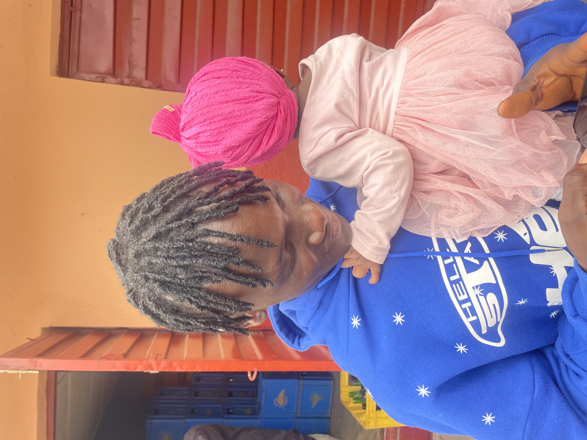 A young Black male with dreadlocks, wearing a blue hoodie with white star and "HELL STAR" graphics, is holding a baby. The baby is wearing a pink turban and a light pink long-sleeved shirt with a pink tulle tutu. The background appears to be an outdoor setting with orange and red walls and a red door slightly ajar. Blue crates are visible in the background, along with a yellow crate containing bottles. The scene is likely in an urban environment.