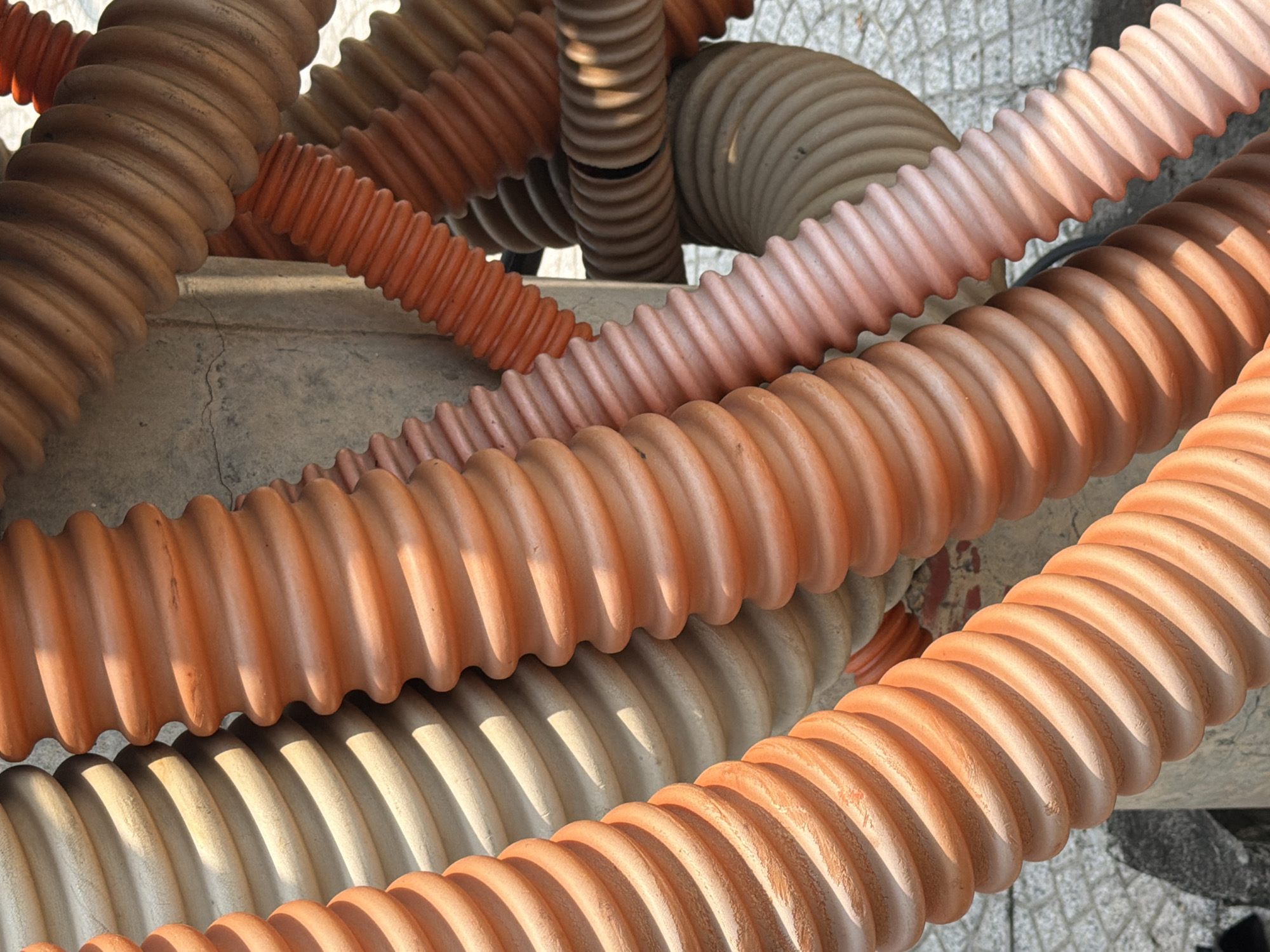 This close-up, high-angle image captures a chaotic arrangement of numerous corrugated pipes or tubes, primarily in shades of orange, brown, light pink, and off-white/beige. The pipes, varying in diameter, are intertwined and overlap, resting on and around a rough, light grey concrete surface. Some pipes appear to be stacked, while others snake across the frame in a seemingly random pattern.
No people are visible, and no specific activity is taking place; the pipes appear to be static, possibly stored, discarded, or part of an unfinished installation. The scene suggests an outdoor setting, potentially a construction site or storage area in Đà Nẵng, Vietnam.
Notable details include the strong, direct sunlight casting distinct, crisp shadows that accentuate the ribbed texture of the corrugated material. This lighting emphasizes the three-dimensional form of each pipe and creates a dynamic interplay of light and shadow across the entire composition. The color palette is dominated by warm, earthy tones, contrasted by the cooler grey of the concrete and the lighter beige/pink pipes. There is no visible text or identifiable branding on any of the pipes. The ground beneath and between some pipes shows hints of pavement or tiles.