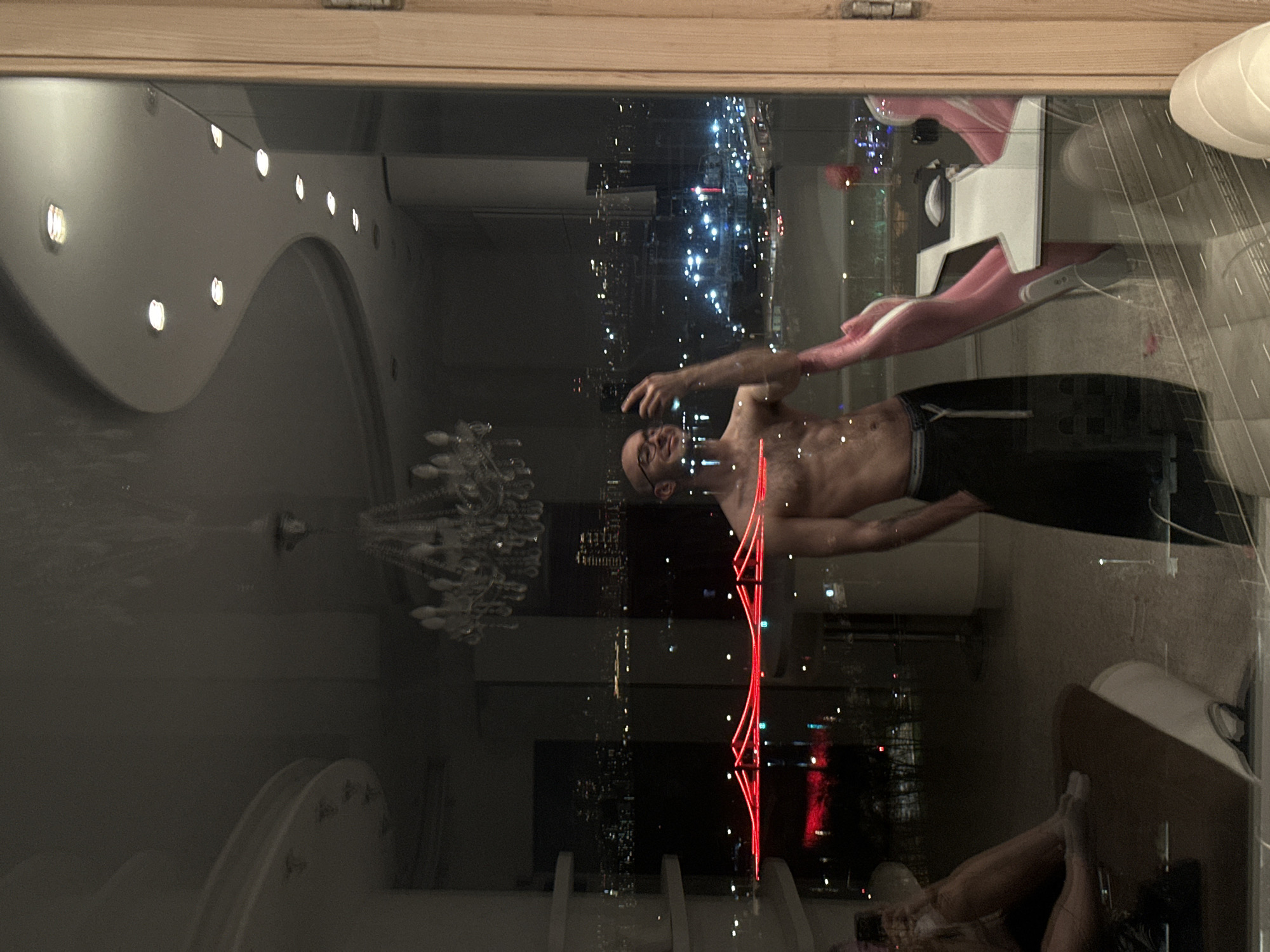 A shirtless man with glasses stands indoors, likely near a window, with a cityscape reflection visible on the glass. The cityscape includes lights suggesting a nighttime setting and potentially tall buildings. Inside, there is a chandelier and a decorative ceiling with lights. Furniture such as chairs and a table are also visible in the reflection. The location is potentially Moscow, Russia.