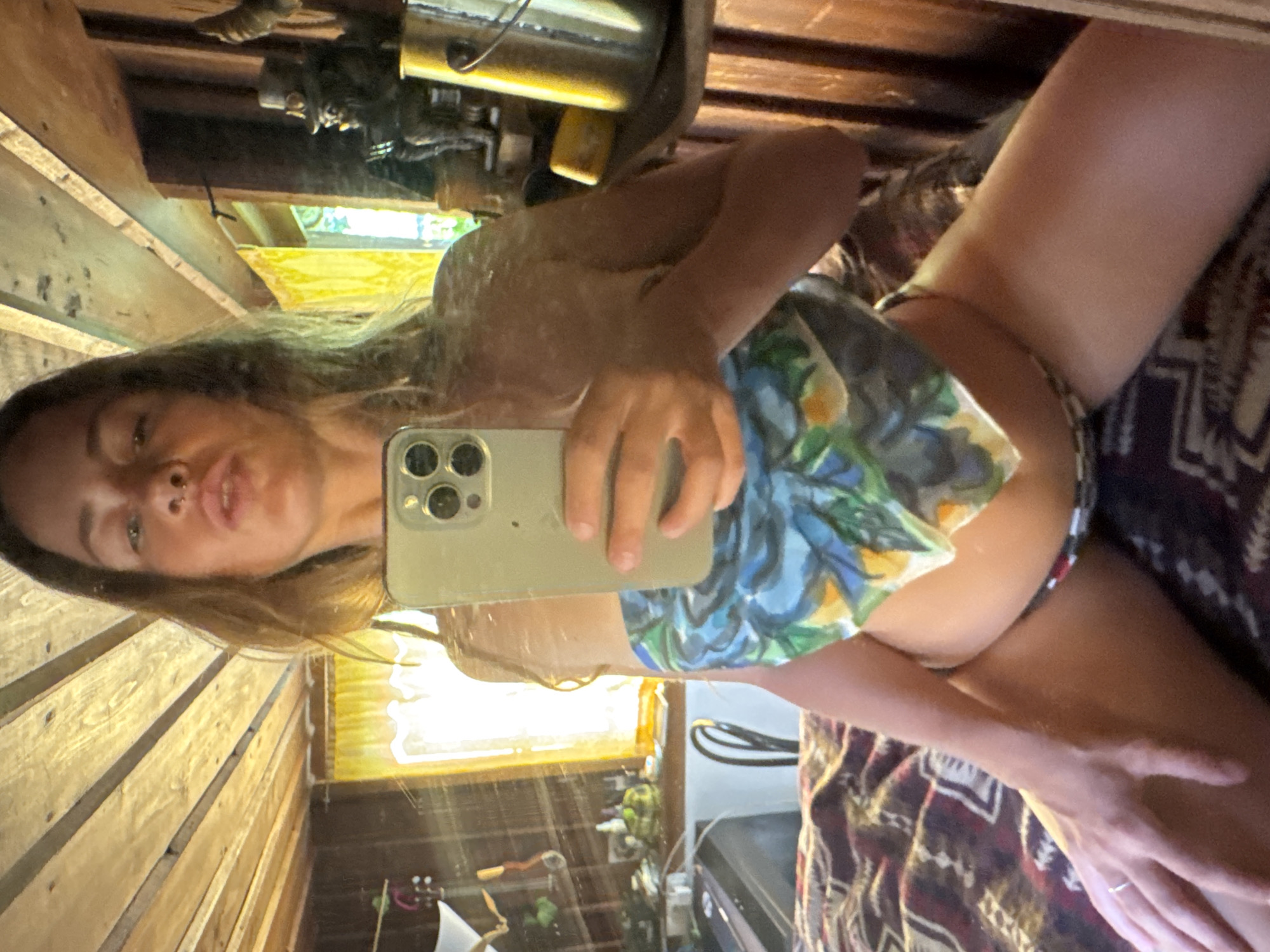 The image is a mirror selfie taken by a person indoors. The person is holding a smartphone and appears to be wearing a floral-patterned top or dress. Their hair is long and brown, and they are looking at the camera with a slight pout. The background suggests a rustic or cabin-like setting, with wooden walls and possibly a window with yellow curtains. There are various objects reflected in the mirror, including what looks like shelves with items on them and some sort of metal appliance. The lighting suggests it is daytime. The overall mood is casual and personal.
