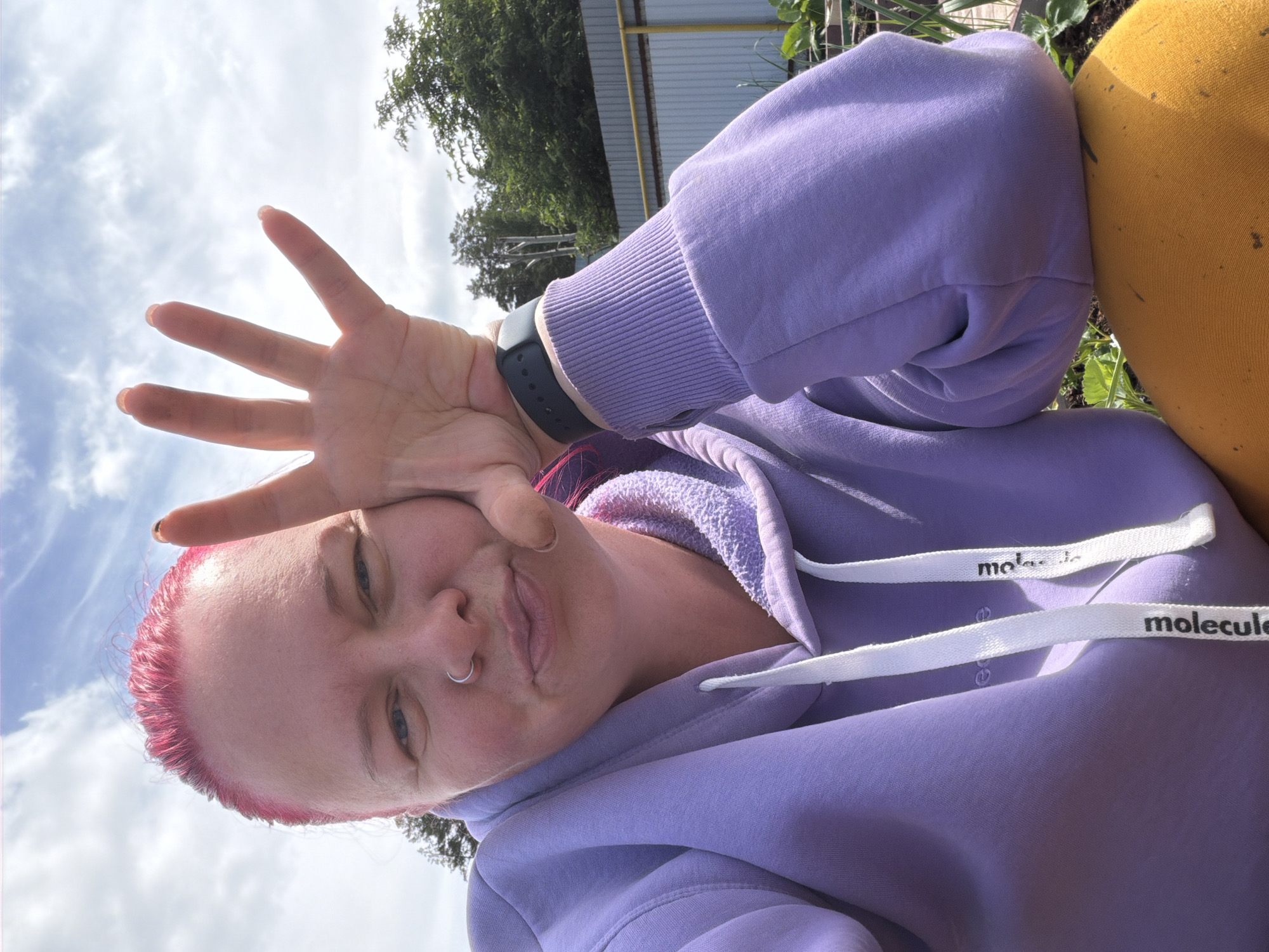 A person with short, vibrant reddish-pink hair, fair skin, and a nose ring in their left nostril is featured in a playful outdoor selfie. They are wearing a light purple hooded sweatshirt (hoodie) with prominent white drawstrings, which clearly display the text "molecule" and a partial "mo" from the same word. A dark-colored fitness tracker or smartwatch is visible on their left wrist.
The person is holding their left hand up to the camera, palm facing forward with fingers spread wide, creating a 'stop' or 'five' gesture that partially covers their face. They are looking towards the camera with a playful, slightly pouting expression.
The setting is outdoors during the daytime under a bright, partly cloudy sky. In the background, green trees and lush foliage are visible, suggesting a natural or semi-natural environment. Behind the trees, a light-colored corrugated metal structure with a yellow horizontal bar can be seen. To the right of the person, a section of yellow fabric, possibly part of another garment or object, is visible. The overall ambiance is casual and lighthearted. This image was captured in Chaikovsky, Russia.