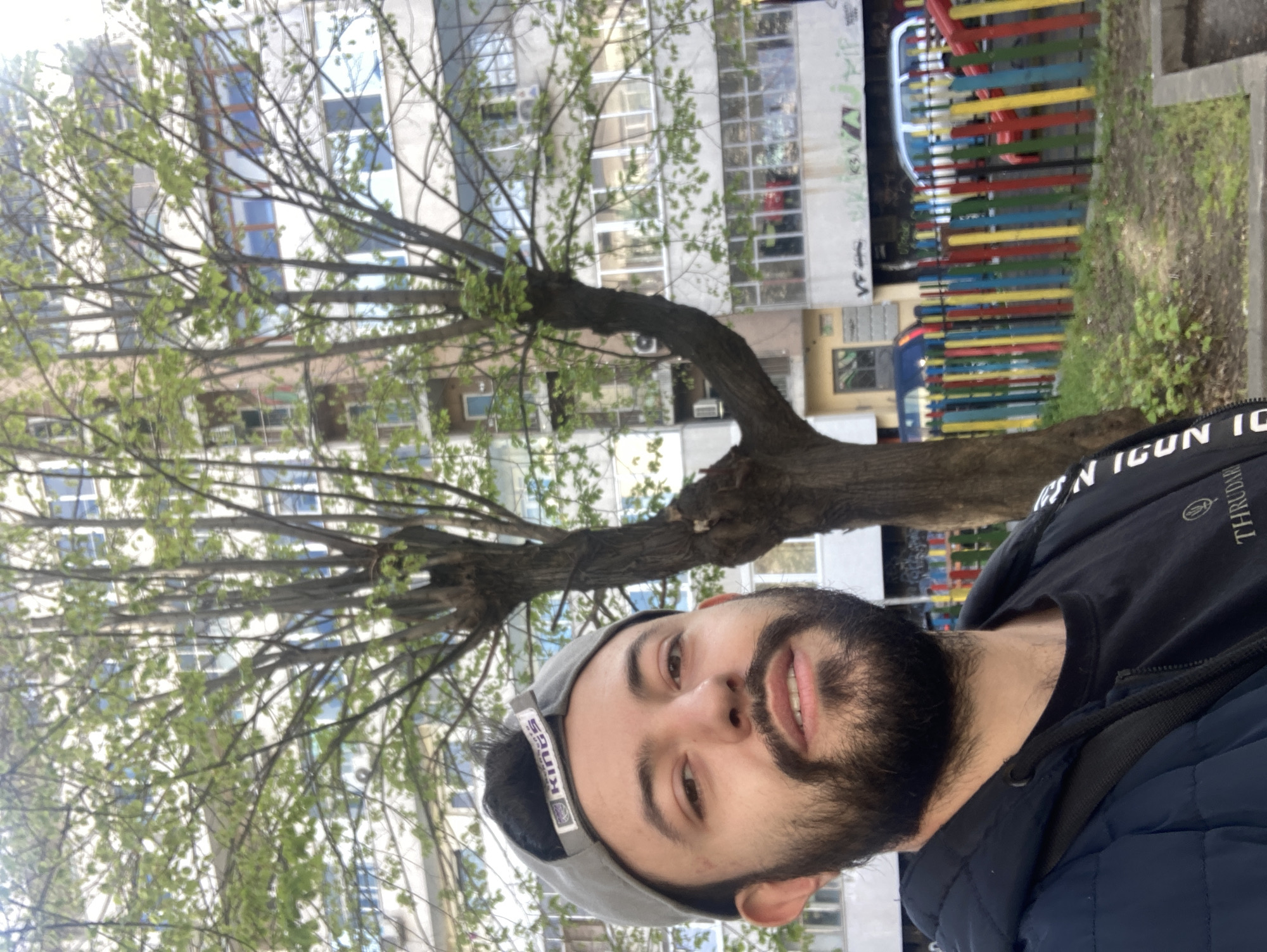 The image is a vertical outdoor portrait, likely a selfie, of a young man in an urban setting, taken during daytime.
The primary subject is a young man with a dark beard and mustache, sporting a slight, pleasant smile as he looks directly at the camera. He wears a grey baseball cap backward, displaying the word "KINGS" on the rear band. His attire includes a black t-shirt with stylized white text, parts of which are legible as "N IGUN ICON IO" and "THRUDAHR," layered under a dark blue puffer jacket. A black strap from a bag is visible over his right shoulder.
The background features a large tree with bare branches and vibrant new green leaves, indicative of springtime. Behind the tree, several multi-story buildings with numerous windows are visible, typical of residential or commercial architecture in an urban environment. To the right, a distinctive fence with vertical slats painted in alternating red, yellow, blue, and green colors suggests a nearby playground or recreational area. Faint, illegible graffiti can be seen on some building walls.
The lighting is soft and diffused, suggesting an overcast or partly cloudy day, or a time of day when the sun is not directly overhead, such as late morning or early afternoon. The overall impression is casual and relaxed. The architectural style and presence of a colorful play area are consistent with a city like Varna, Bulgaria.