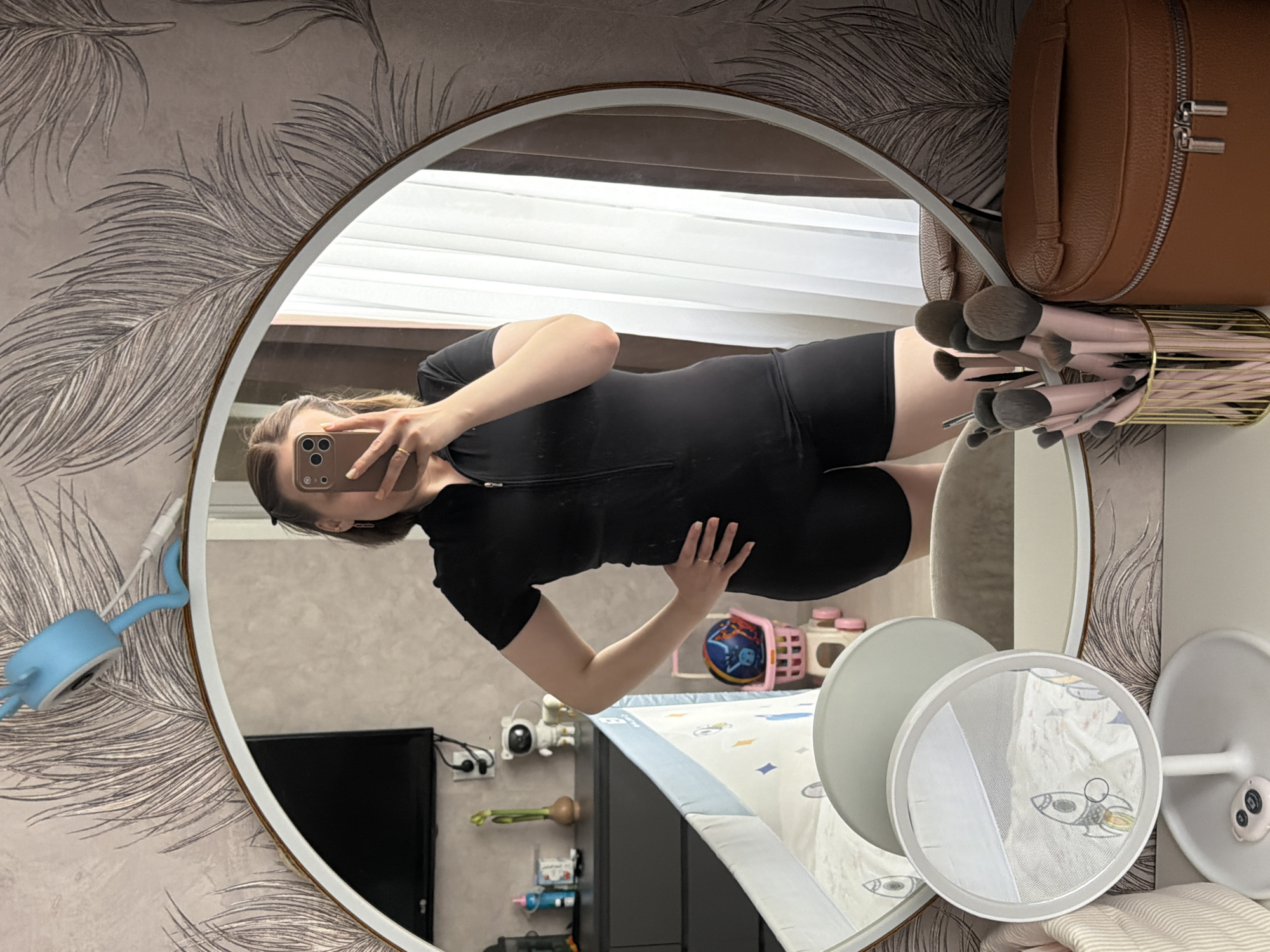A young woman is captured in a mirror selfie, standing in what appears to be a bedroom or nursery in Reshetnikovo, Russia. The image is presented sideways, with the room's features reflected in a large, round mirror framed in gold.
The woman, with light brown hair, is wearing a fitted black, short-sleeved bodysuit or shapewear, zipped down the front. She holds a smartphone with a tan case, partially obscuring her face, as she takes the photo. Her left hand rests on her lower back, and a ring is visible on her left ring finger.
The room's decor includes wallpaper adorned with large, stylized grey feather patterns. Reflected in the mirror are various items indicating a home setting, possibly a child's room. These include a bed or crib with a sheet patterned with rockets and stars, a white circular light or baby monitor on a stand, and a pink basket containing a toy ball. Other reflections show a black television screen, power outlets with cables, a small white astronaut figurine, and a blue flexible light or toy with a cartoonish head. A gold-colored container filled with makeup brushes featuring pink handles and dark bristles is visible on a surface next to a brown leather-like bag with silver zippers. The overall lighting suggests it is daytime. No visible text is discernible in the image.