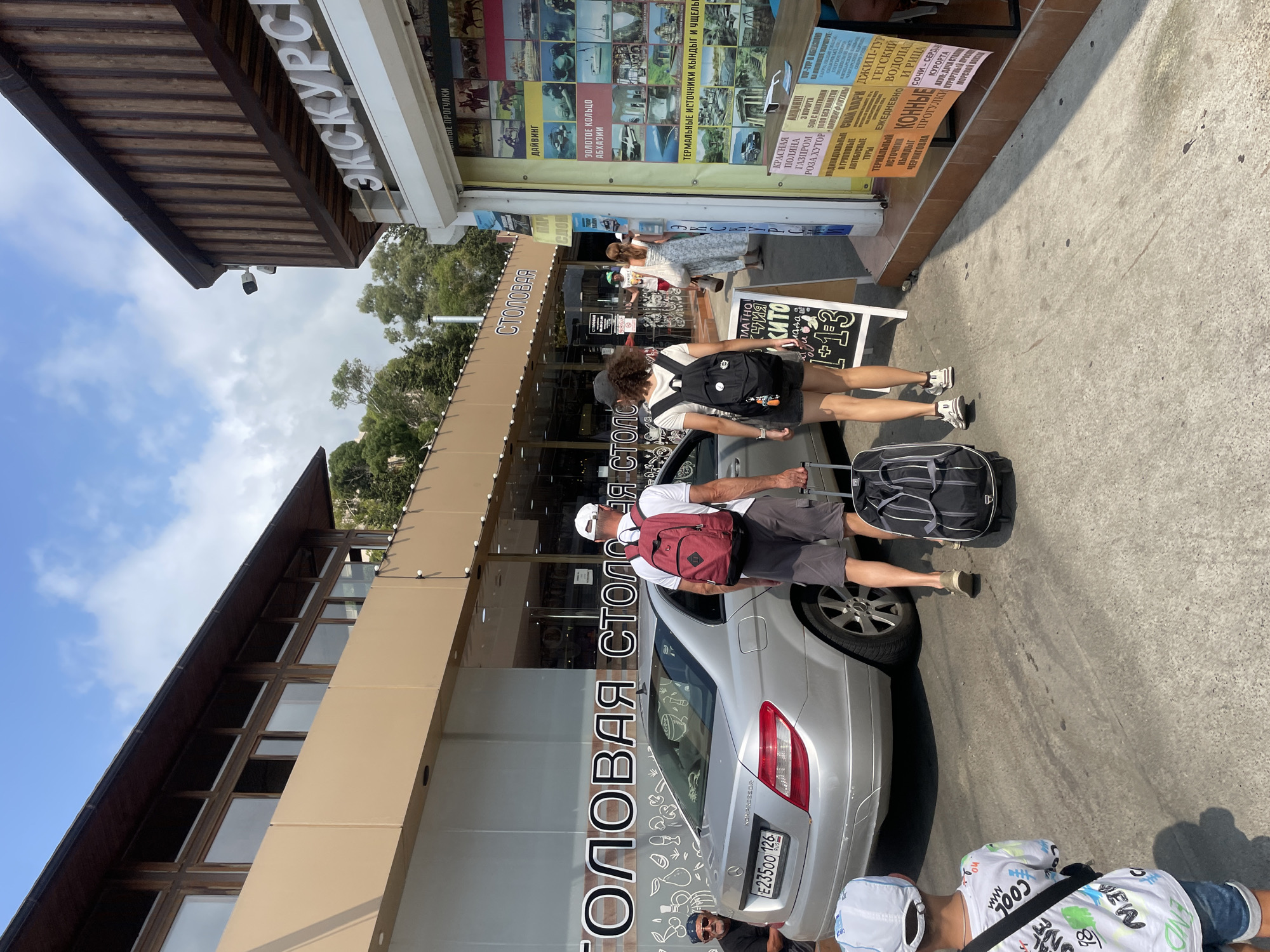 This is a full-shot, eye-level, outdoor, daytime photograph of a street scene in Sochi, Russia. In the foreground, a young boy wearing a white t-shirt with colorful writing and blue denim shorts is facing away from the camera, looking towards a silver Mercedes-Benz C-Class sedan. Behind the boy, a man and a woman, both wearing casual clothing and backpacks, are walking away from the camera towards a building with a sign that reads "СТОЛОВАЯ" (dining hall). The man is pulling a black rolling suitcase. To the right of the couple is a kiosk with a sign that reads "ЭКСКУРСИИ" (excursions), displaying advertisements for various tours and activities. The building on the left has large windows and a wooden exterior, while the kiosk on the right is brightly lit and covered in posters. The street is paved and appears to be an urban environment with trees visible in the background. The sky is blue with scattered clouds.