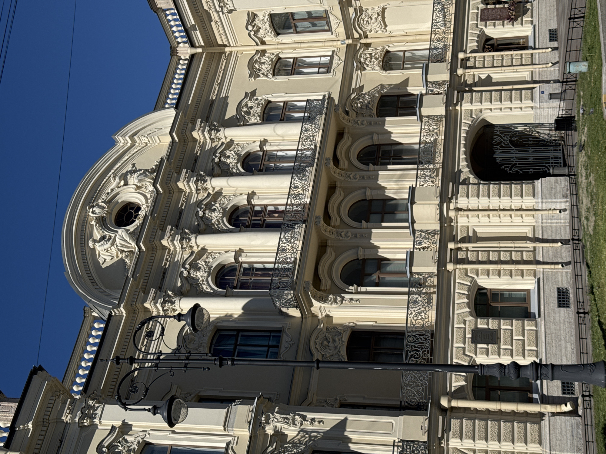 This image depicts the exterior of the Stroganov Palace, a prominent 18th-century Baroque/Rococo building located on Nevsky Prospekt in Saint Petersburg, Russia. The multi-story palace facade is painted in a pale cream or yellow hue, under a clear, bright blue sky with thin overhead power lines.
The central section of the palace features four engaged columns supporting a curvilinear pediment. Within the pediment is a circular oculus window, surmounted by a reclining sculpted figure holding a wreath, flanked by elaborate stucco ornamentation. Below the pediment, the second floor displays three large arched windows, fronted by a continuous, ornate wrought-iron balcony. Additional arched windows, some with individual smaller balconies, are visible on the upper floors and extending wings.
The ground floor is characterized by rusticated stone blocks, with arched window openings and a large central arched entrance featuring a dark, decorative wrought-iron gate. Several light-colored vertical drainpipes are affixed to the facade. A rectangular, dark historical plaque is mounted on the rusticated wall to the right of the main entrance.
In the immediate foreground, a tall, black, ornate cast-iron street lamp with two globes stands to the left. A strip of green grass, bordered by a low black metal fence, separates the viewer from the building. Several pigeons are scattered across the grass. A black waste bin is situated near the palace's main arched entrance, and a light blue/green bollard or container is visible on the grass further to the right. Bright sunlight casts distinct shadows, emphasizing the building's architectural textures and relief details.