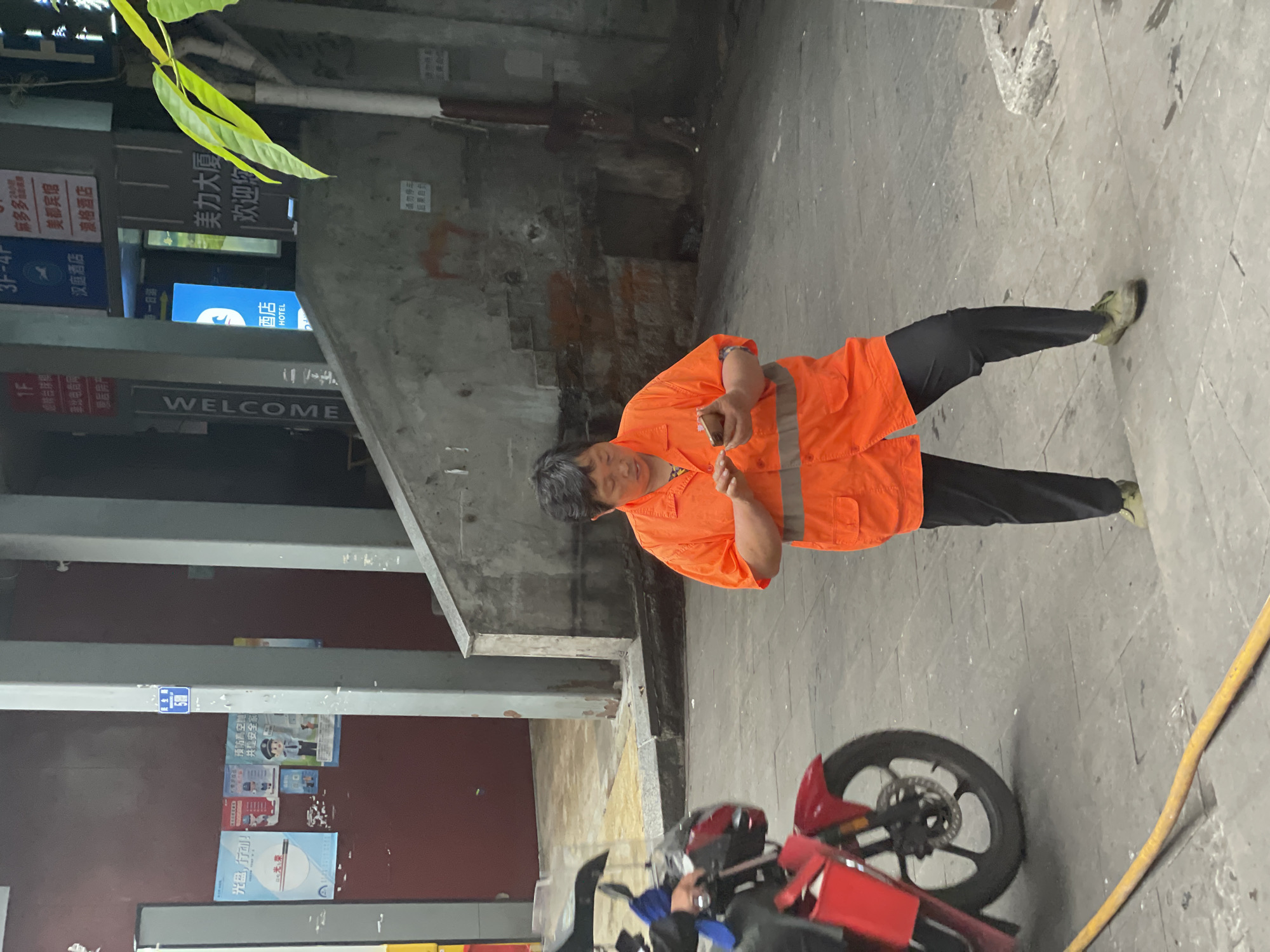 The image shows a woman wearing an orange safety vest and dark pants standing on a paved surface. She appears to be looking at or interacting with a smartphone in her hands. To the right of the woman is the back wheel of a red motorcycle parked. The surrounding environment includes urban elements such as a building façade with signage, including "WELCOME" and hotel advertisements. Graffiti and posters can be seen on the walls. The location can be assumed to be in the city of Yuzhong District, China, as implied by the text on the storefronts.