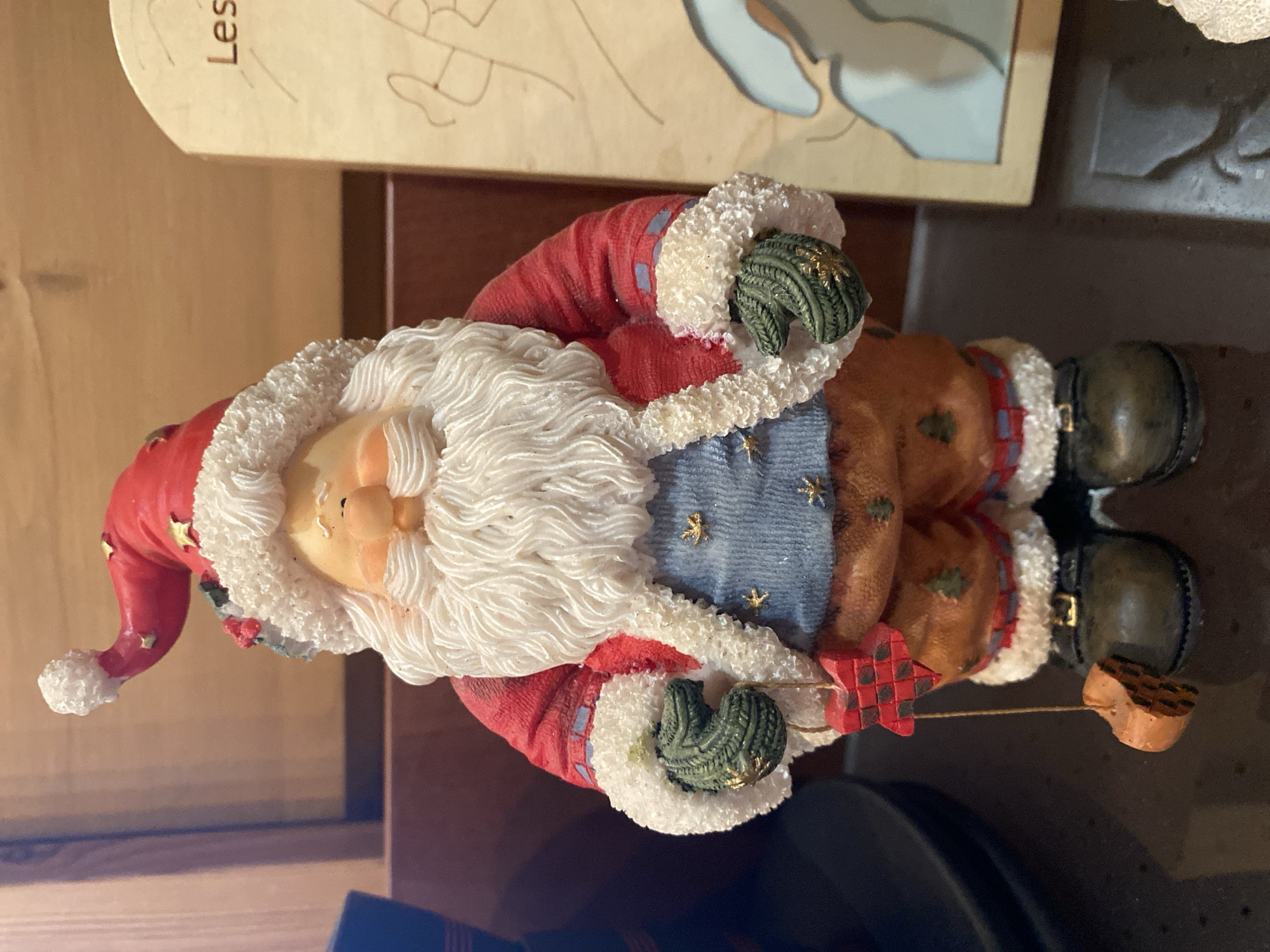 A vertically oriented, medium shot photograph features a figurine of Santa Claus displayed indoors. The figurine is detailed, with a red suit trimmed in white fur, a blue vest adorned with gold stars, and brown pants. Santa has a white beard and rosy cheeks, and he is holding a red and black checkered star. His hands are gloved in green. Behind the figurine, a light-colored object with the Cyrillic text "Лес" is visible. The background suggests an indoor setting, possibly a shelf or table, with a dark surface in the foreground and a wooden surface behind. The lighting appears to be artificial, and the overall mood is festive and decorative.