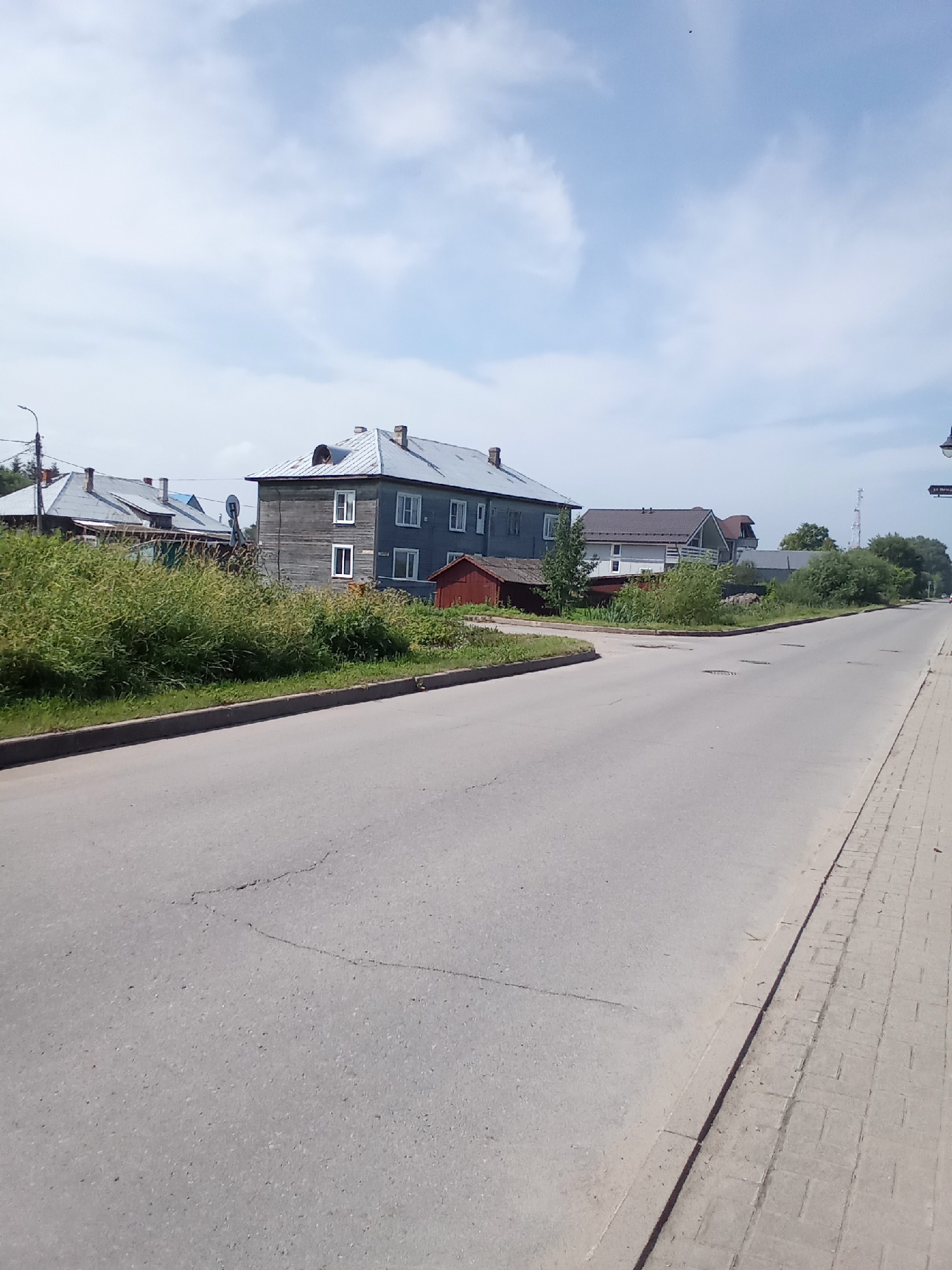 The image captures a view of a residential street in Belozersk, Russia, on a clear day with scattered clouds. The street, paved with asphalt, curves slightly to the left. To the left of the street, a thick patch of green bushes lines a curb, separating it from the road. Behind the bushes, several houses are visible. The most prominent is a two-story wooden house with a grey facade and a metal roof. It features multiple windows with white frames. Adjacent to this house, to the left, is another house with a similar grey, pitched roof and some visible wooden siding. A small, red, shed-like structure is situated in front of the main house. Further back, other houses with pitched roofs and some greenery can be seen. On the right side of the street, a paved sidewalk made of rectangular blocks runs parallel to the road. A lamppost is visible on the far right side of the image, and a tall, lattice tower, possibly a communication tower, stands in the background to the right of the houses. The scene depicts a quiet, suburban or small-town environment.