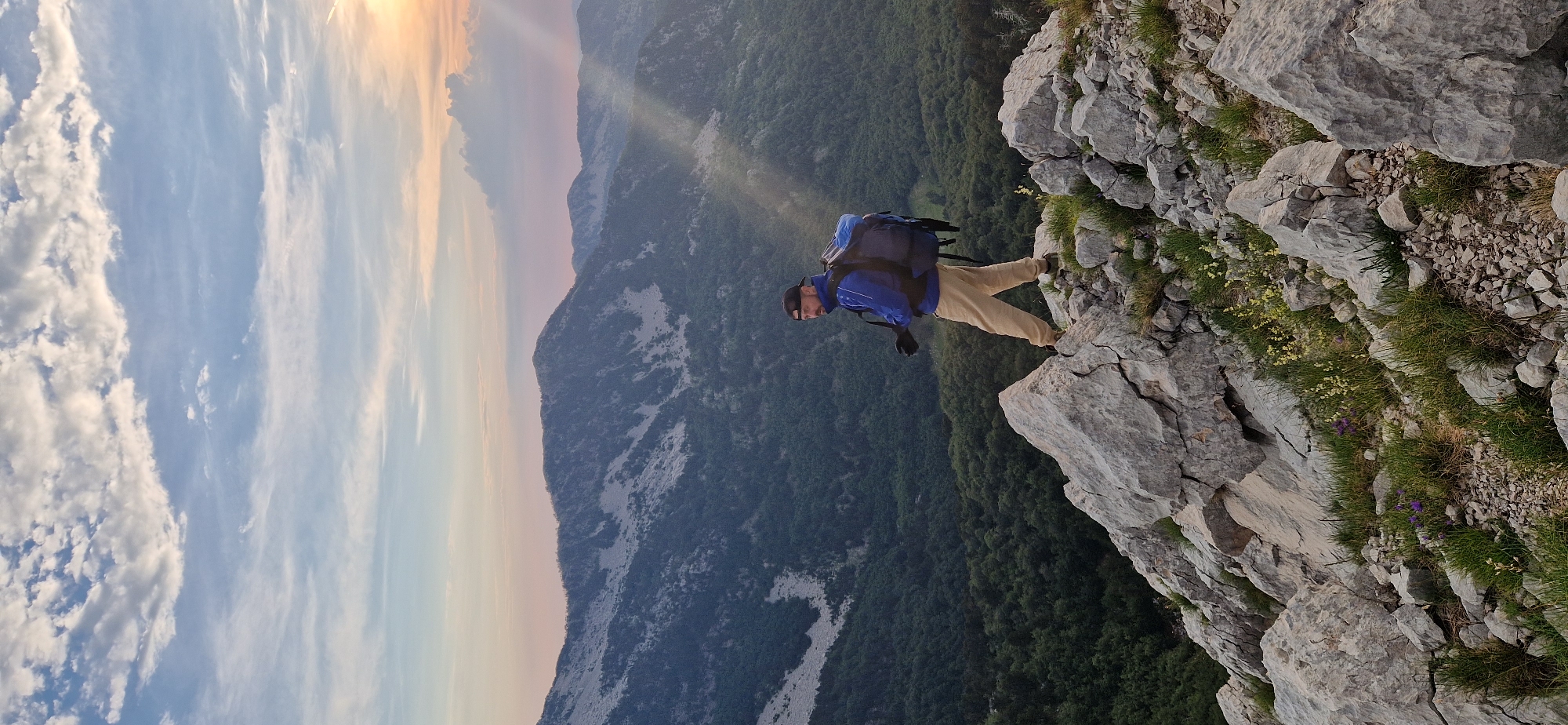 A man wearing a blue jacket, tan pants, a black baseball cap, and a large backpack stands on a rocky mountain peak. He is facing to his left, looking out over a vast valley filled with green trees. In the background, more mountains are visible, with a colorful sunset sky above. The foreground is dominated by large, gray rocks and scattered green grass with small purple and yellow wildflowers. The location is identified as Ubla, Bosnia and Herzegovina.