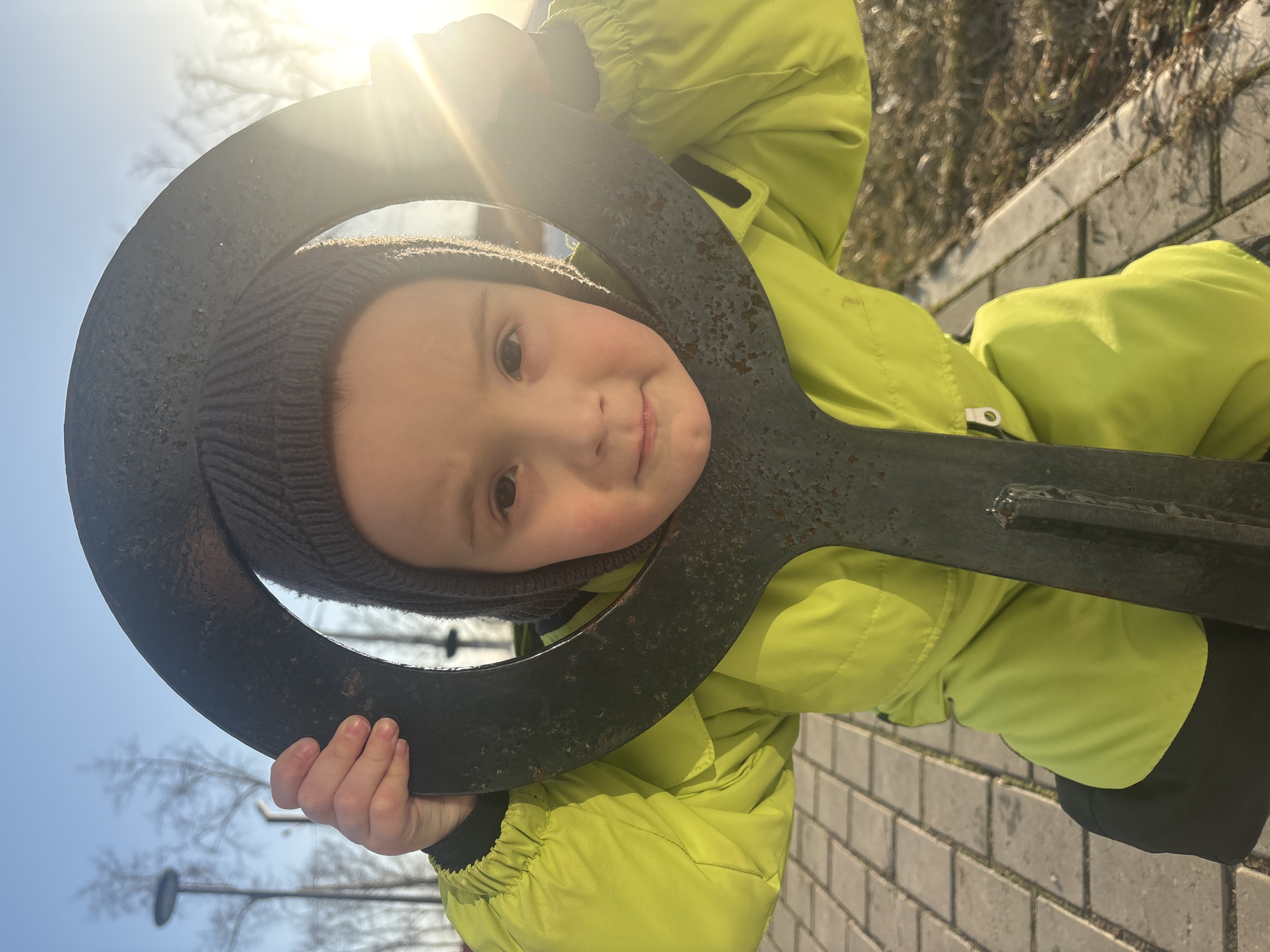 A young child is depicted outdoors in Chaikovsky, Russia, looking directly at the viewer. The child wears a lime green padded winter jacket and a dark grey knitted beanie. Both hands grasp a dark, textured, O-shaped object, which frames the child's head and upper torso. The child's face is illuminated by direct sunlight, prominent on the right side. The ground beneath the child consists of light-colored rectangular paving stones. The background features a clear blue sky, several bare deciduous trees, and a street light pole with a circular fixture on the left. Bright sunlight from the upper right generates a visible lens flare. To the child's right, the paved area gives way to dry, sparse vegetation bordered by a concrete curb.