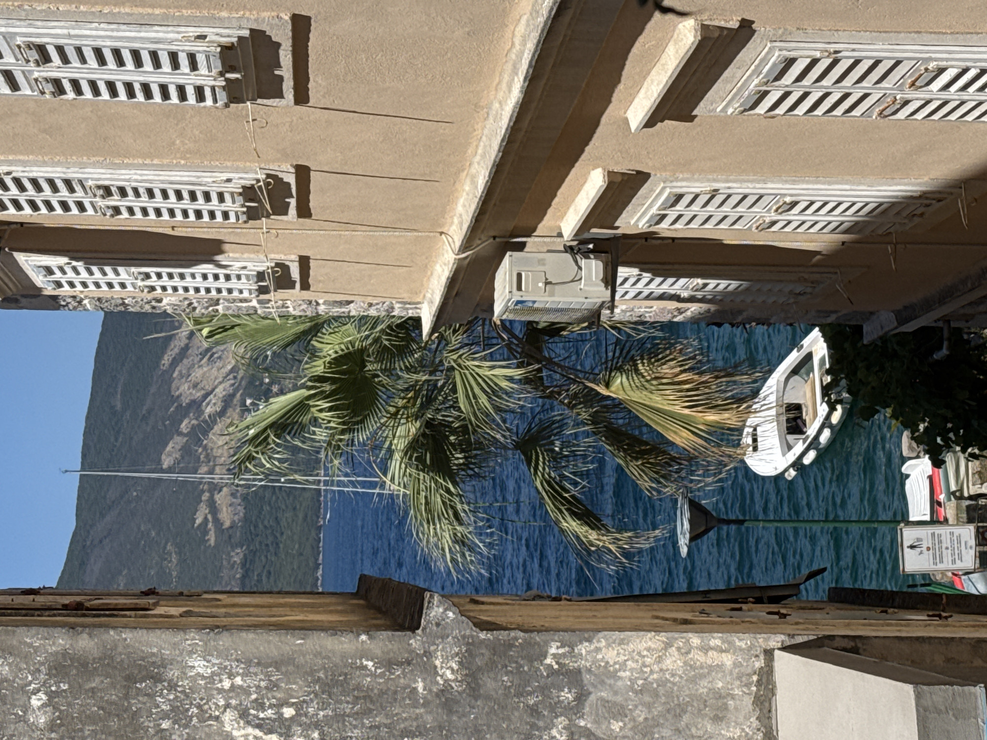 The image shows a narrow alleyway with buildings on either side, opening onto a view of the sea and a mountain. The building on the right is light brown with several windows covered by white shutters. An air conditioning unit is mounted on the exterior wall of this building. The building on the left is weathered and gray, with a section of its facade appearing to be peeling.
In the background, the blue sea is visible, with a white sailboat anchored in the water. A small white boat is also floating near the shore. A palm tree with lush green fronds partially obscures the view of the sailboat. In the distance, a verdant mountain rises under a clear blue sky.
At the bottom of the alley, there is a street lamp and a sign with pictograms indicating rules about dogs and possibly littering. Several other items, possibly chairs or small boats, are stacked near the sign. The overall scene suggests a Mediterranean coastal town.