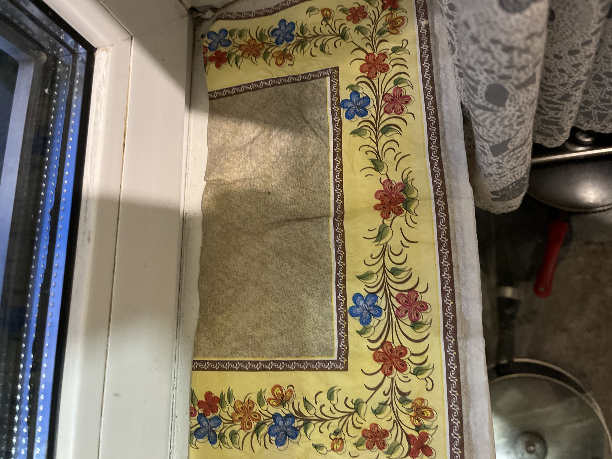A close-up, high-angle, indoor shot shows a yellow fabric with a floral design placed on a windowsill in Minsk, Belarus. The fabric has a patterned border and a plain central area. Below the windowsill, part of a metal pot and a red handle are visible. The scene is lit by ambient light.