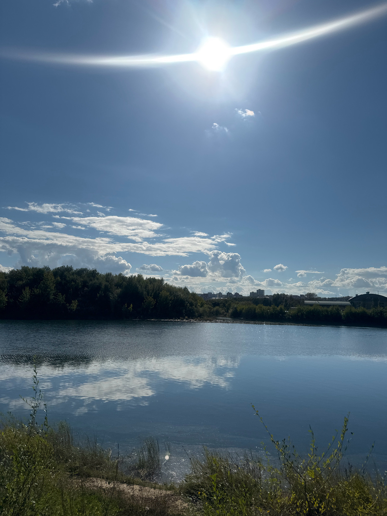 A wide shot captures a tranquil lake scene under a bright, sunny sky with a few scattered clouds. The sun is high and strong, casting a bright glare across the upper portion of the frame. The lake's surface is calm, reflecting the sky and the dense line of trees that form the backdrop. Beyond the trees, a cityscape is visible in the distance, characterized by buildings and structures. The foreground shows a bank covered in grass and some brush. The scene is located on a body of water in Irkutsk, Russia.