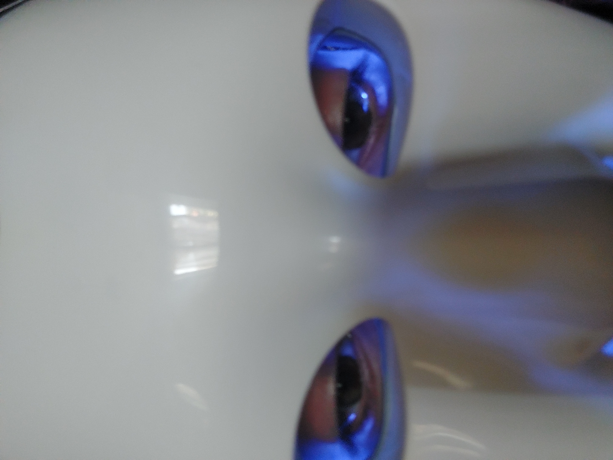 A close-up, frontal photographic capture depicts the upper portion of a smooth, glossy white surface resembling a stylized human face or mask. Two distinct eye apertures reveal dark human eyes, which are prominently illuminated by an intense blue light originating from within the eye sockets. This blue light casts a vibrant hue onto the surrounding contours of the white material and reflects in the pupils. Above the eye line, two distinct, rectangular white light reflections are visible on the highly polished forehead area, indicating overhead artificial light sources. A subtle, rounded protrusion suggesting a nose bridge is centrally positioned below the eye openings. The overall impression is that of a humanoid robot or a person wearing a futuristic mask. The scene is located in the city of Unknown, Unknown.