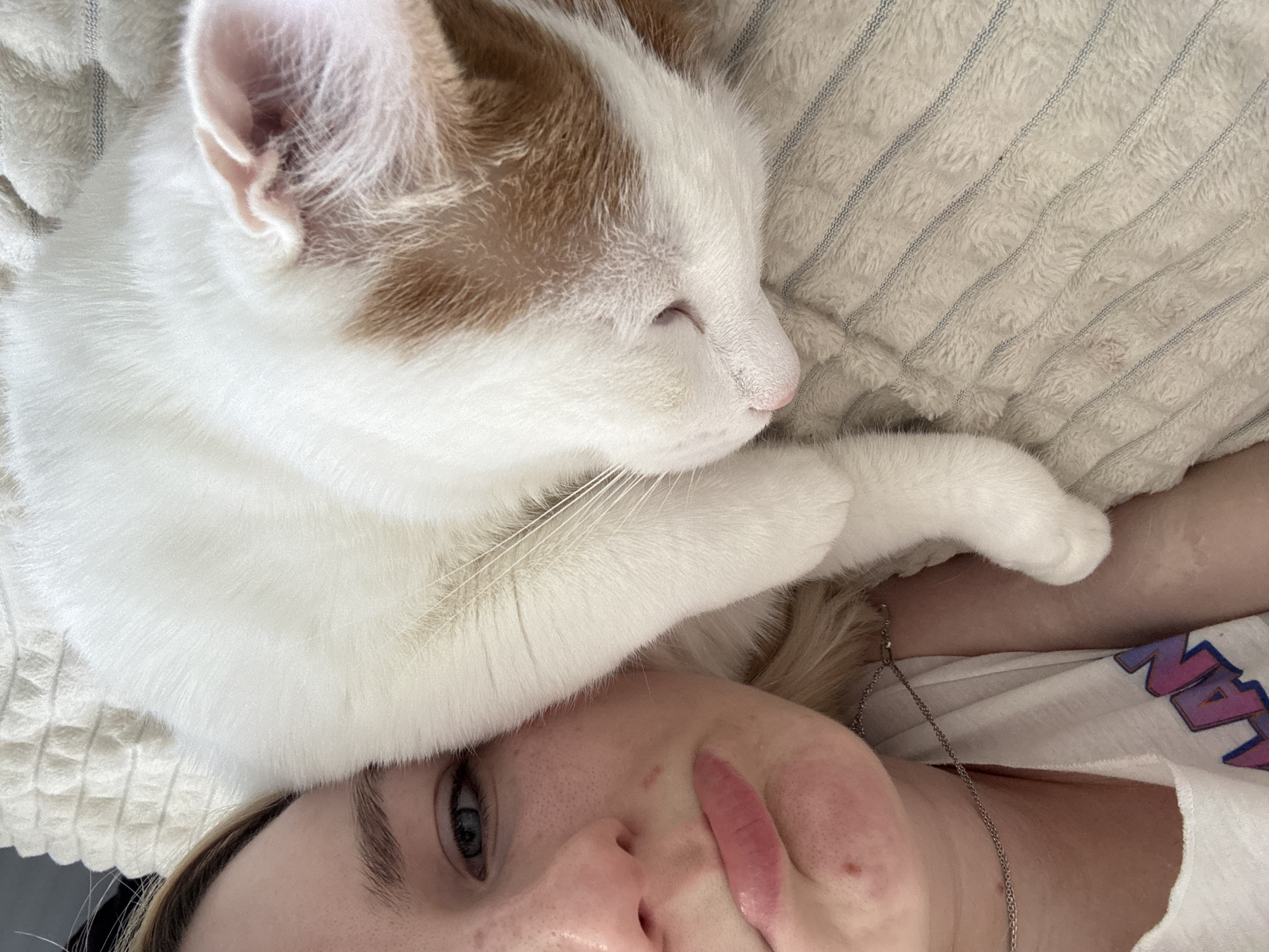 A person is lying down with a white cat with orange patches sleeping on their head. The person has fair skin, freckles, and light blonde hair. They are wearing a white t-shirt and a silver chain necklace. The cat is mostly white with orange patches on its head. They are lying on a light beige pillow with faint striped texture.
There is no information available to determine whether the photo was taken in Vsevolozhsk, Russia or any other location. The provided image does not contain any location-specific visual clues or metadata.