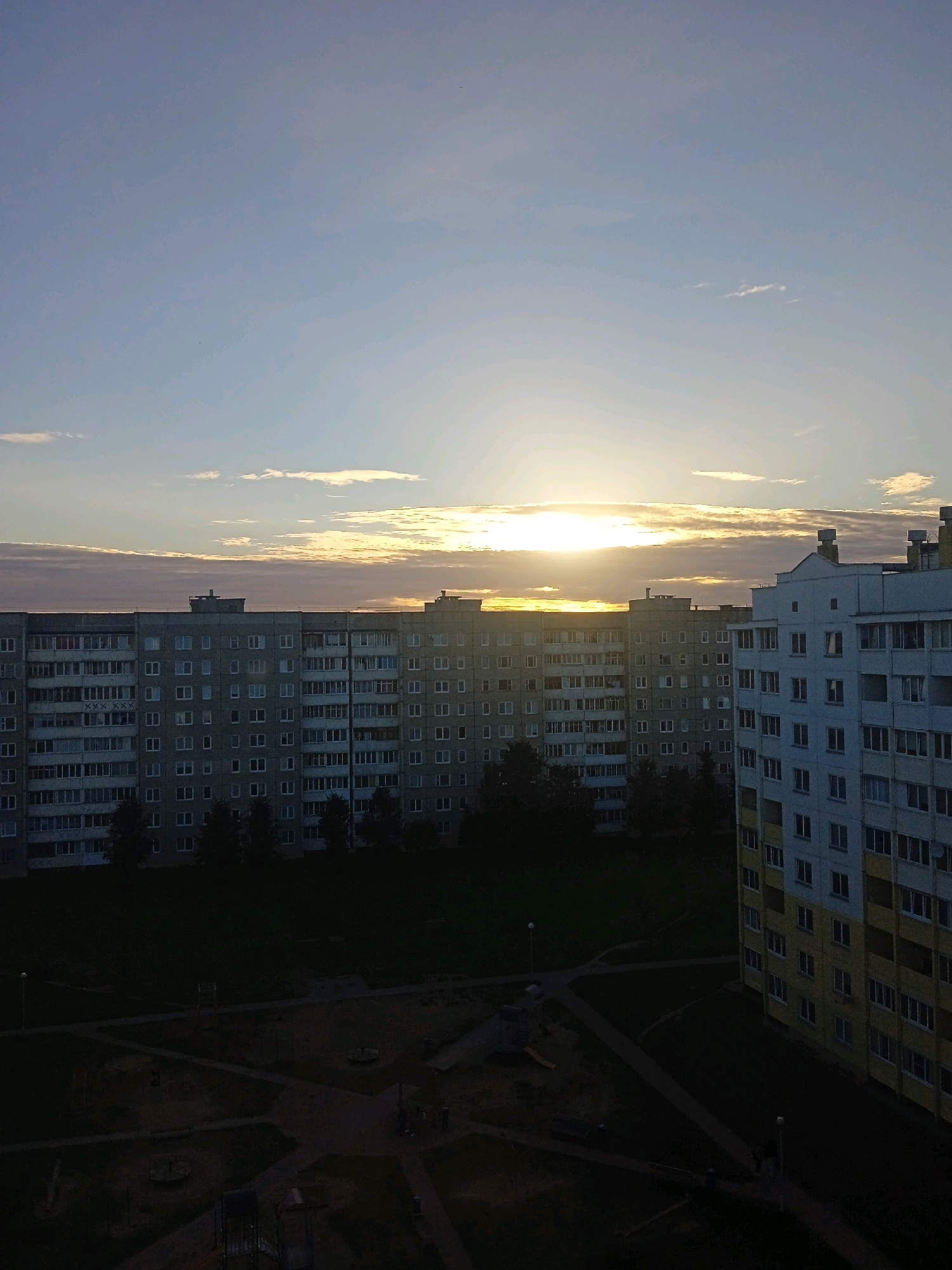 The image depicts a cityscape at dusk, likely in Lida, Belarus, characterized by a cluster of Soviet-era apartment buildings. The sky is a gradient of pale blue and grey, with the setting sun casting a warm, golden light through scattered clouds. The sun, a bright, intense orb, is positioned near the horizon, partially obscured by the long, horizontal buildings. These structures, appearing to be of uniform height and design, stretch across the frame, their facades featuring numerous windows. One building on the right is brighter, with a yellow and white exterior, contrasting with the grey of the others. In the foreground, the scene transitions to a darker, more shadowy area depicting a courtyard or playground. This area is sparsely populated, with a few indistinct figures walking on pathways. There are playground structures visible, suggesting a residential area. The overall impression is one of a quiet, possibly residential, evening scene in a post-Soviet urban environment.