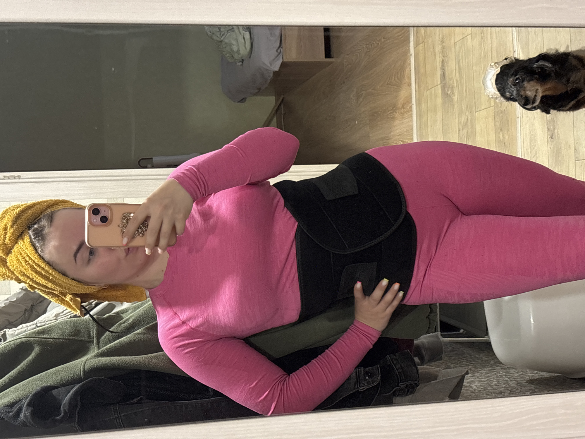 A person is captured in a mirror selfie, standing in what appears to be a bedroom or dressing area. They are wearing a bright pink long-sleeved athletic top and matching leggings, complemented by a black waist trainer or support belt around their midsection. A yellow towel is wrapped around their head, suggesting a recent shower or hair treatment. The individual holds a light-colored smartphone, possibly an iPhone, with a decorative grip, partially obscuring their face. Their fingernails are painted with colorful tips in shades of yellow, orange, and white.
The reflection in the mirror reveals various details of the room. Part of the floor is covered in light wooden planks, while another section has a grey patterned carpet. Piles of clothes, including a green fleece jacket and dark denim jeans, are visible on the left side of the reflection. A white plastic basin or tub rests on the carpeted floor. In the upper right corner of the mirror, a small, dark-furred dog with a white/grey muzzle looks upwards. Wooden furniture, possibly shelves or a cabinet, is also reflected in the background. A window or covered opening appears dark in the upper left of the reflection. The scene portrays a casual, domestic moment in Izhevsk, Russia.