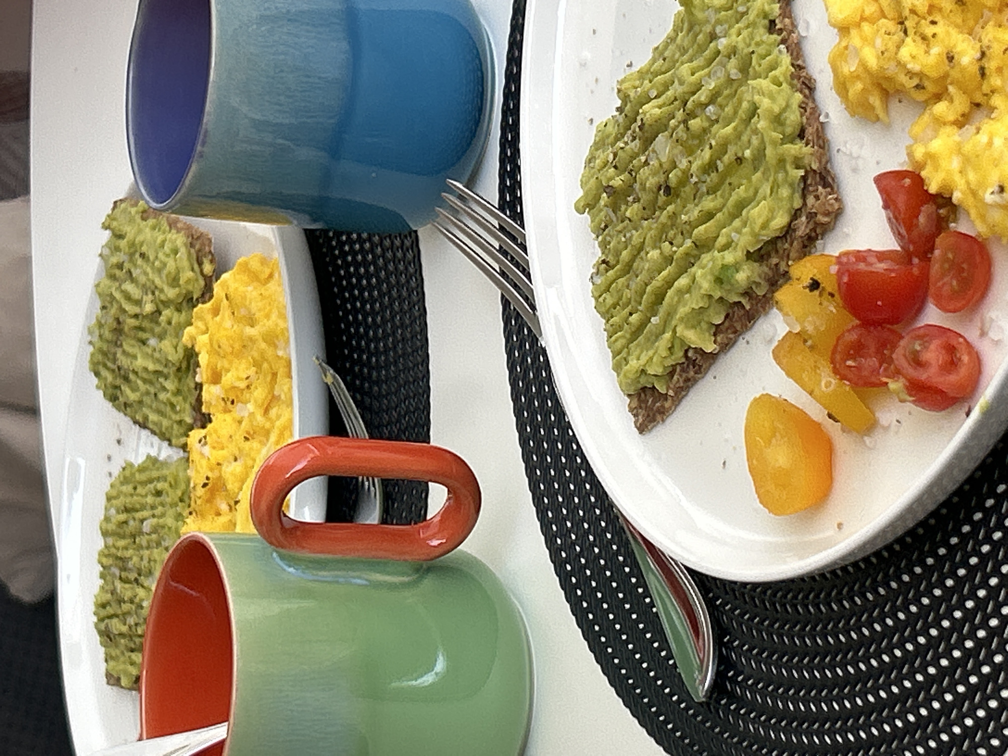Two plates of breakfast are laid out on a white surface, partially covered by black placemats with a white dotted pattern. Each plate contains scrambled eggs, mashed avocado on toast, and cherry tomatoes. One plate also has a few pieces of yellow bell pepper. A blue mug is tilted over one plate, and a green mug with an orange handle is positioned near the other. A fork is visible between the blue mug and its plate. The food appears fresh and well-seasoned, with visible salt flakes. The lighting suggests it is daytime, likely morning, and the setting appears to be indoors, possibly a home or a cafe. There are no people visible in the image, but the presence of food and cutlery indicates a meal is being prepared or consumed. The scene is static, with no discernible activity beyond the arrangement of the food. The location context of Cupra Marittima, Italy, does not provide any specific visual cues within the image itself.
