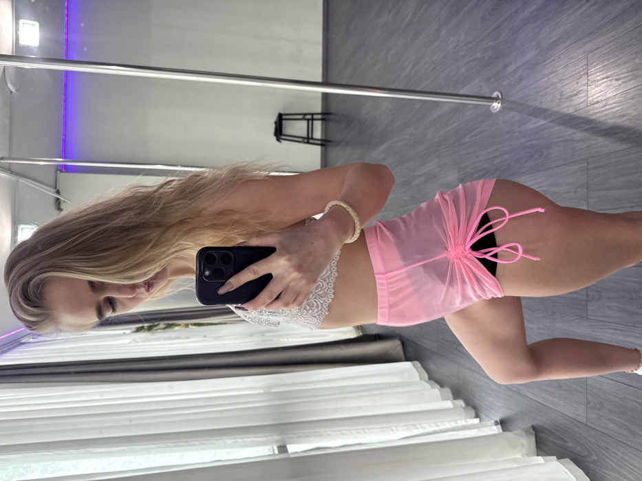 A blonde female subject is depicted in a mirror selfie taken within what appears to be a pole fitness studio in Unknown, Unknown. The subject, positioned slightly to the left of center, holds a black smartphone in her right hand, angled towards her reflection. She is wearing a white lace bralette and sheer pink mesh mini shorts with adjustable drawstrings over black undergarments. A light-colored, possibly woven or beaded bracelet adorns her right wrist. Her long, wavy blonde hair cascades over her shoulders. The studio features a dark grey floor, light-colored walls, and a chrome-colored pole extending vertically from floor to ceiling on the right side of the frame. A dark, three-legged stool is visible in the background reflection on the right. White sheer curtains and a darker grey curtain are present on the left side of the image, suggesting a window. A strip of purple LED lighting is visible along the upper right wall in the reflection.