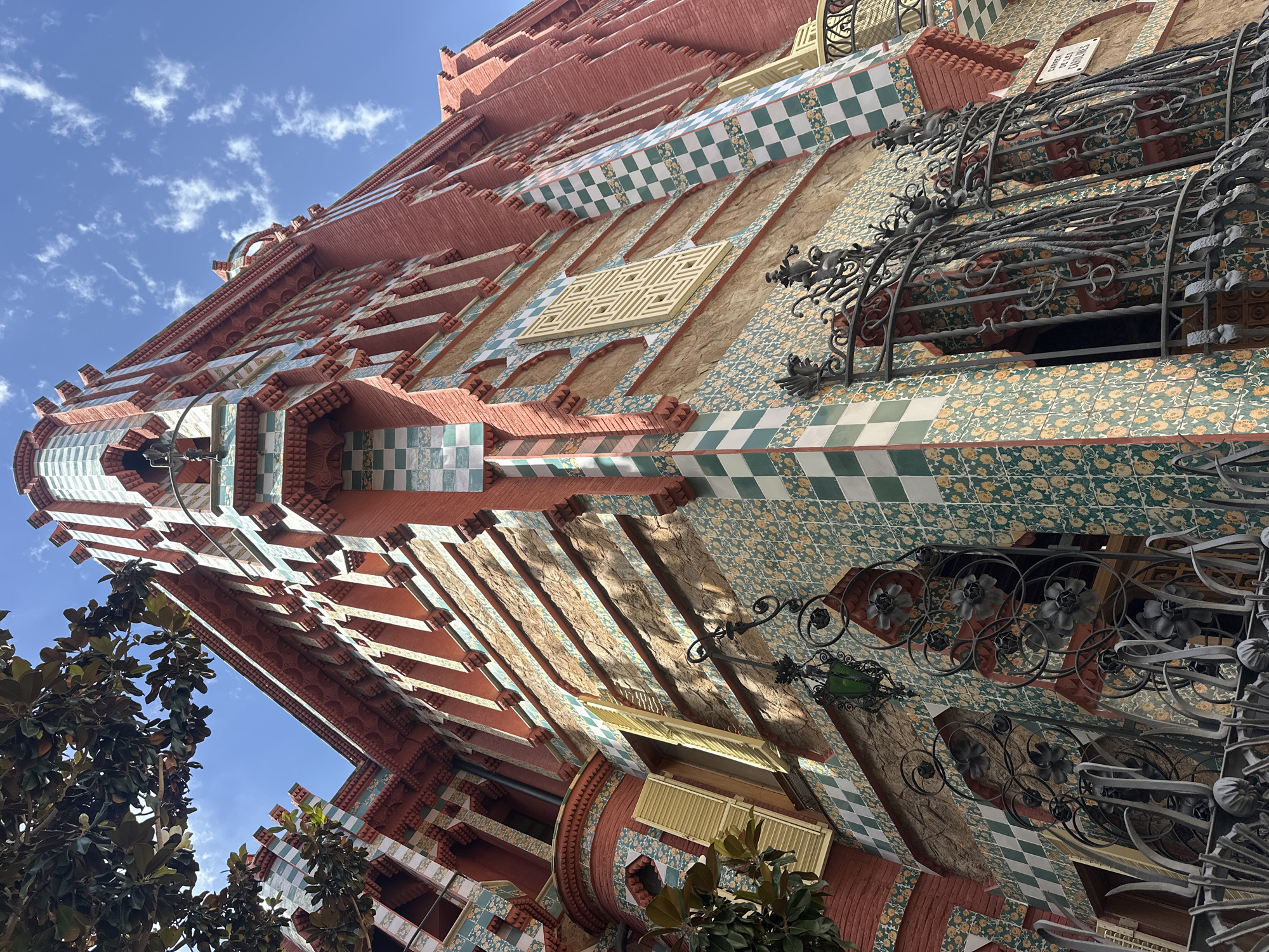 The image shows the exterior of Casa Vicens, located in Barcelona, Spain. It is a building with a reddish-pink brick facade, adorned with intricate decorative tiles in green, white, and yellow. The architecture features sharp angles, arched windows with green shutters, and ornate wrought-iron balconies. A tree with large, dark green leaves is visible in the upper left corner, casting shadows on the building. The sky above is a clear blue with scattered white clouds. The image is a low-angle shot, emphasizing the height and elaborate details of the building.