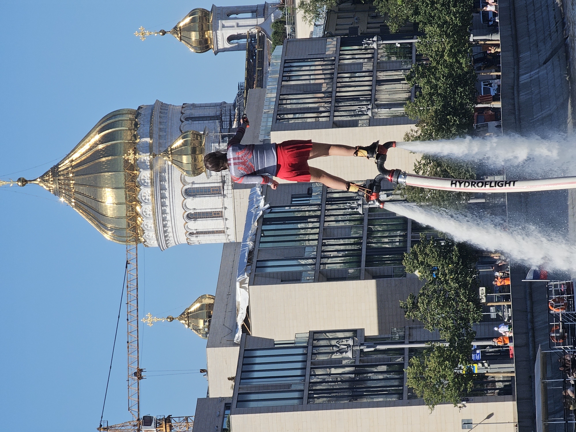 An individual is performing on a hydroflight device above a body of water, with a city skyline in the background. The person, seen from behind, is wearing a grey long-sleeved shirt with red accents and a red skirt. They are standing on a device that sprays water upwards, propelling them into the air. In the background, there is a large building with a golden dome, characteristic of Russian Orthodox churches. The building appears to be under construction or renovation, as evidenced by a crane visible to the left of the frame and scaffolding on parts of the structure. Adjacent to this building is a modern, multi-story building with reflective glass windows. Trees line the waterfront, and a boat with passengers is visible in the foreground. The sky is clear and blue. The overall scene captures a recreational activity set against a backdrop of historical and modern architecture in Moscow, Russia.