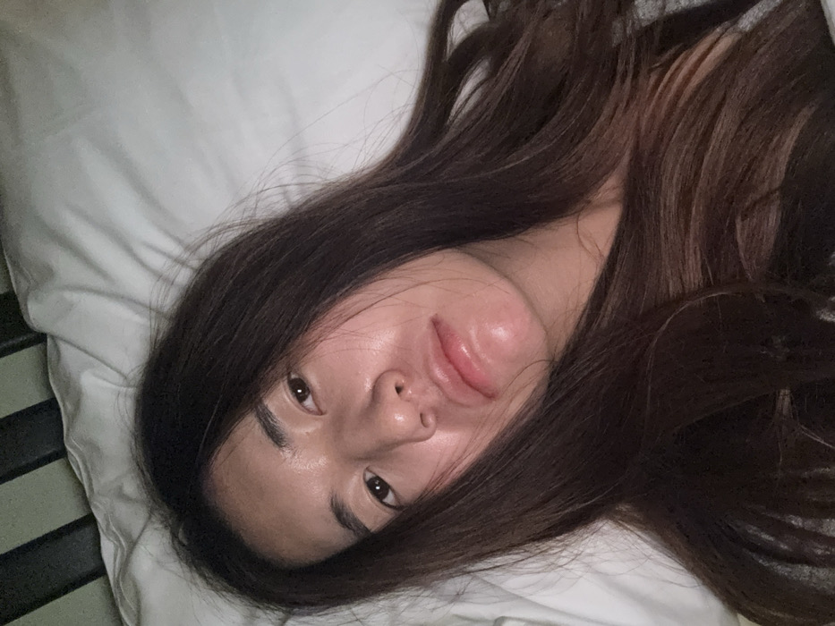 A close-up, high-angle portrait shows a young adult female, appearing to be of East Asian descent, lying down indoors. Her long, dark brown hair frames her face, falling across her shoulders and chest. She has a fair complexion with a slight reddish tone on her cheeks and chin, and her lips are full, slightly parted, and glossy. Her dark eyes are open, looking directly towards the camera, and her expression is neutral, possibly appearing drowsy or subtly pouting. She is resting on a white pillow, which fills the right side and upper portions of the background. A dark, vertical element, consistent with a bed frame or headboard, is partially visible along the left edge of the frame. A small portion of a grey garment is visible on her lower right side. The lighting is soft and diffuse. No visual information within the image permits the identification of its location in the city of Le Havre, France, or any other specific geographical area.