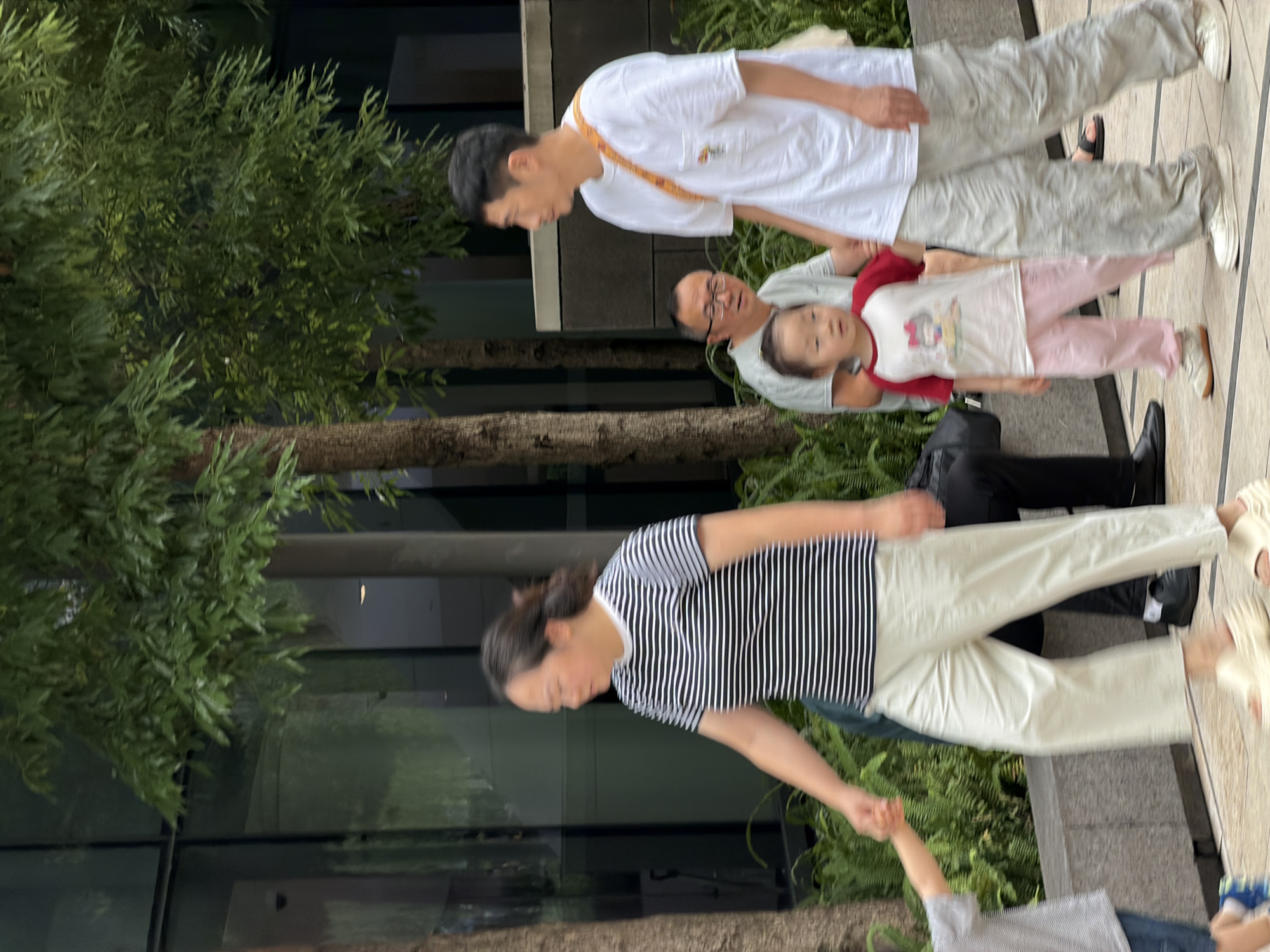In Yuzhong District, China, a family walks along a paved path bordered by trees and a modern building with glass windows. A woman in a striped shirt and light-colored pants walks ahead, holding hands with a child wearing a gray shirt and blue pants. Behind them, a man in a white t-shirt and loose-fitting pants walks with a young girl in a red and white shirt and pink pants. Another man wearing glasses is visible in the background, seated and looking towards the child. The environment appears to be an urban outdoor setting, possibly a park or pedestrian walkway, with lush green foliage and contemporary architecture.