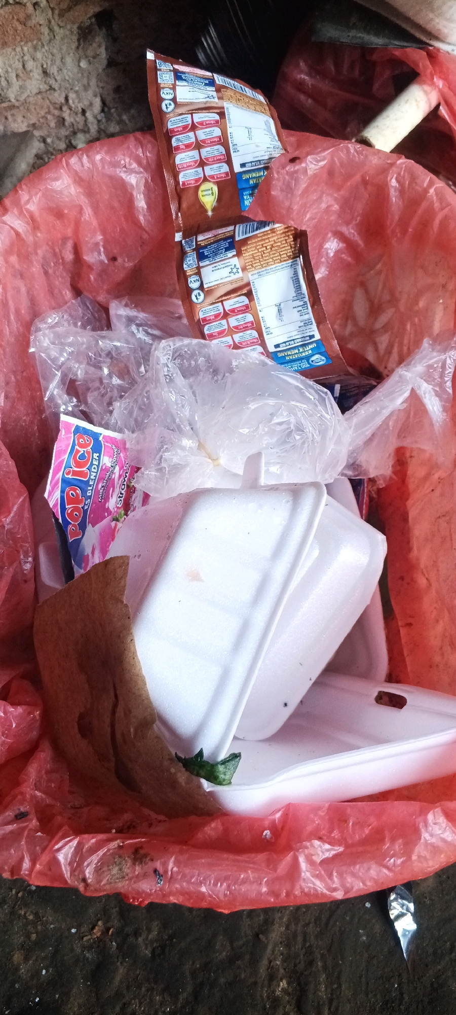 The image displays a close-up, overhead view of a red plastic trash bag filled with various discarded items. Located in Penanggungan, Indonesia, the scene is outdoors on a dirt surface. The contents of the bag include crumpled clear plastic wrappers, a brown food packaging wrapper with text in what appears to be Malay or Indonesian, and several white styrofoam containers, some stacked. One styrofoam container has a small green leaf or vegetable piece on its rim. A pink packet with "Pop Ice" and other text is also visible. The trash bag itself is translucent red, and some of the contents are visible through it. The background shows a rough, textured surface, likely a wall or ground, with some debris. The lighting suggests daytime conditions.
