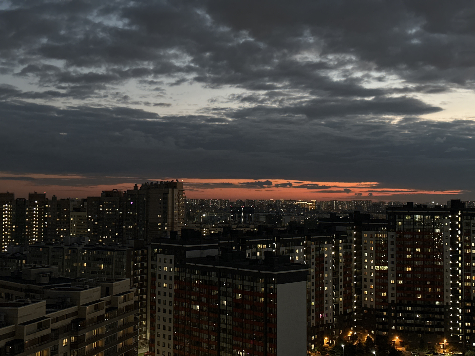 This is an eye-level, wide-angle shot of a densely populated urban area at dusk. The foreground is dominated by a cluster of tall, modern apartment buildings, their facades a mix of white, grey, and red. Numerous windows are illuminated, casting a warm glow against the darkening sky. The architecture appears to be repetitive, with balconies and window patterns forming a rhythmic grid.
In the mid-ground, a sprawling cityscape stretches towards the horizon. This area also consists of numerous buildings, many of which are lower in height than those in the foreground, but still densely packed. Lights are visible throughout this section, suggesting a vast residential or commercial district.
The sky above is dramatic and filled with dark, heavy clouds. A band of intense orange and red light is visible near the horizon, indicating a sunset or the lingering glow of twilight. The overall impression is one of a large, vibrant city at the transition from day to night, with a sense of constant activity suggested by the illuminated windows. The absence of visible people or specific landmarks makes pinpointing a precise location within Russia impossible, but the style of the buildings suggests a modern, post-Soviet era development.