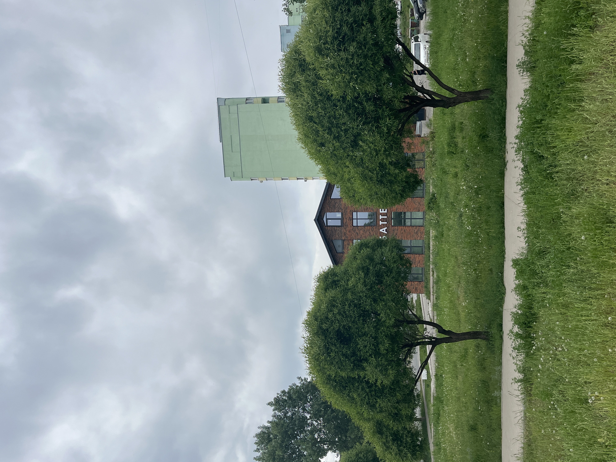 A street scene in Unknown, Unknown features a sidewalk bordering a green area with two trees that partially obscure a brick building with the word "LATTE" vertically written on it. Behind the brick building, a tall green building is visible against a cloudy sky. The grass in the green area appears overgrown. A sidewalk runs along the side of the street. Several parked cars can be seen further in the distance. A few bare trees can be seen.