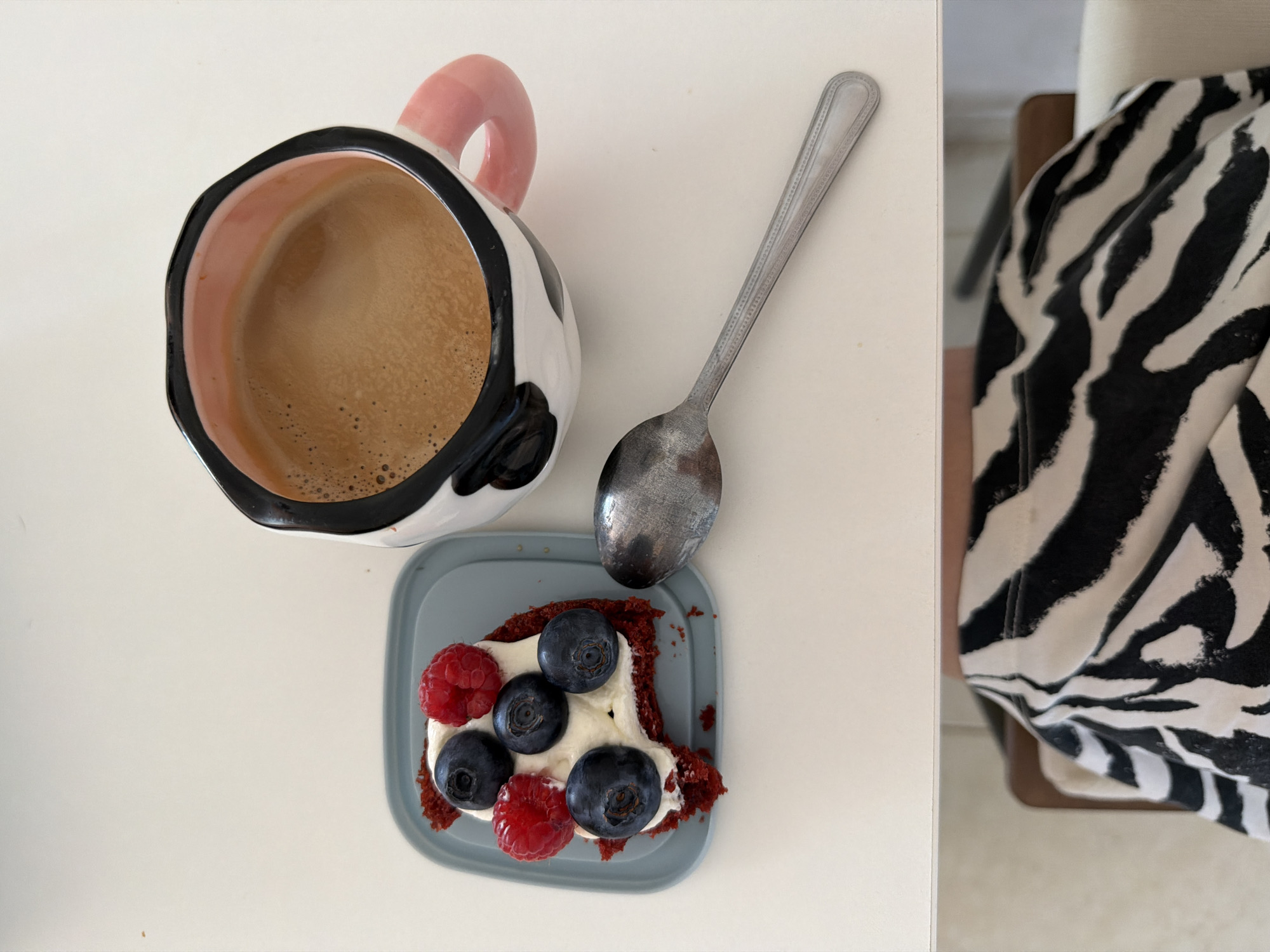 A top-down view shows a white table surface. On the table, there is a cup filled with coffee, a spoon, and a small, square plate holding a slice of red velvet cake. The cake is topped with a dollop of white cream, raspberries, and blueberries. The mug has a black and white design, with a pink handle. A spoon rests next to the plate, pointing towards the top of the image. To the right, a person wearing a black and white zebra-print outfit is visible. The setting appears to be indoors. There is no indication of Tel Aviv, Israel.