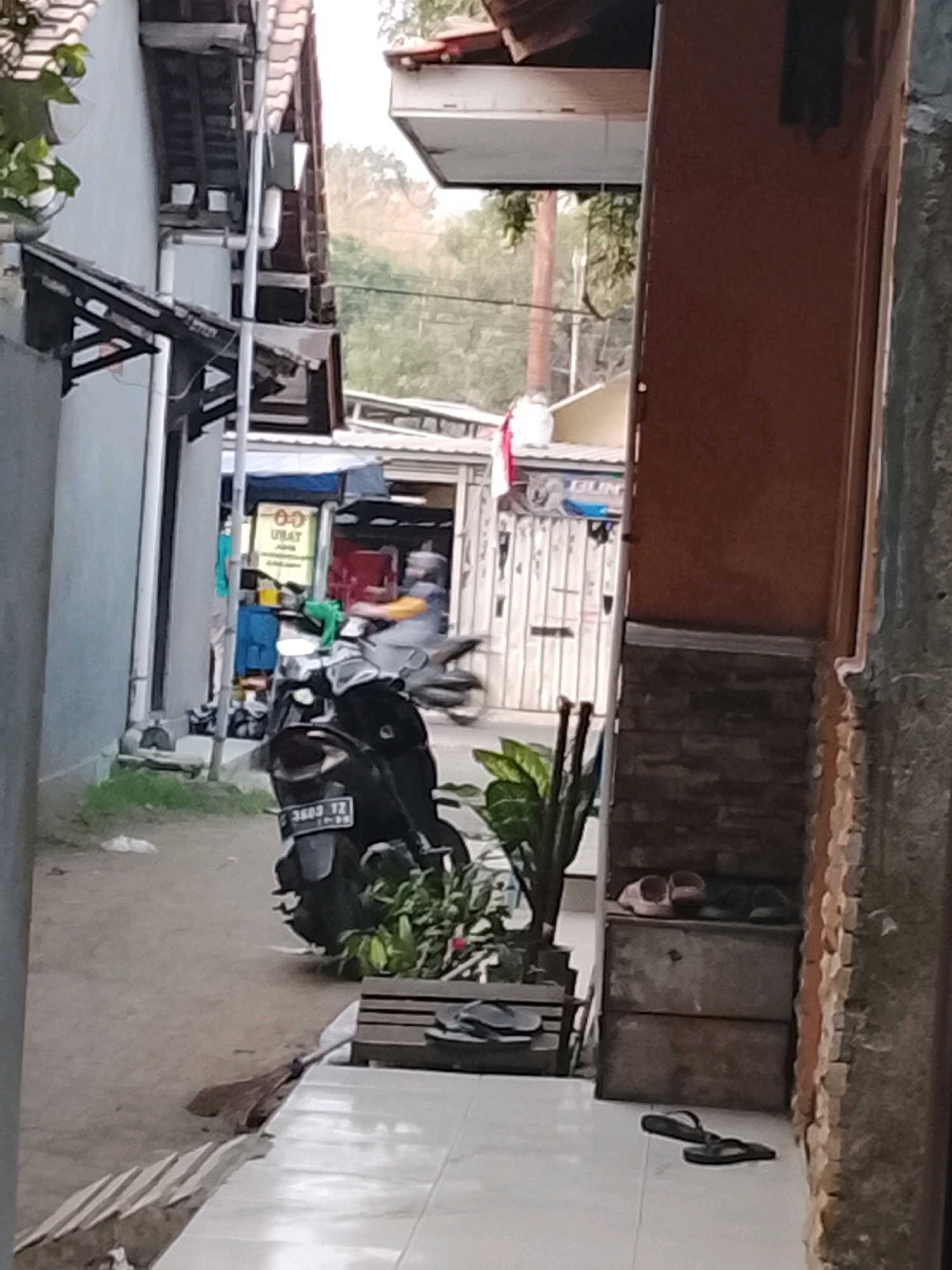The image depicts a narrow alleyway in Ujungrusi, Indonesia. In the foreground, a dark-colored motorcycle is parked, with its license plate visible. A broom lies on the ground near the motorcycle. To the right of the motorcycle, a small wooden structure is visible, topped with two pairs of sandals. Further down the alley, a person is riding a motorcycle, their face obscured by a helmet and the motion blur. The buildings lining the alley appear to be simple structures, some with corrugated roofs and walls made of various materials including brick and light-colored siding. A sign with the word "UBAT" (medicine in Indonesian) is partially visible on one of the buildings. The overall environment suggests a residential or commercial area with limited space. The lighting indicates it is daytime.