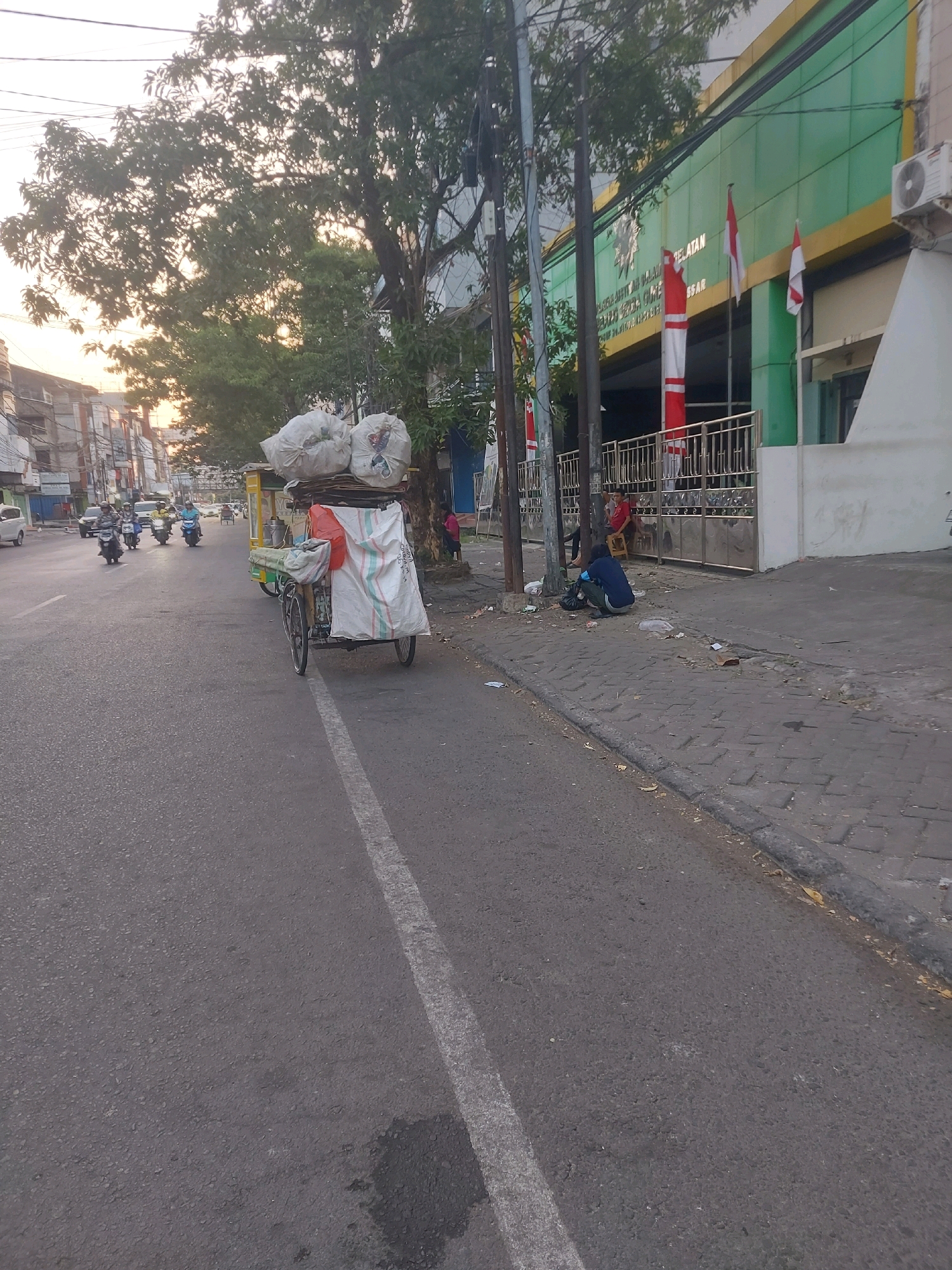 The image depicts a street scene in Makassar, Indonesia, likely during the late afternoon or early morning due to the warm lighting and long shadows. In the foreground, a street with a white dashed line runs diagonally. A pedicab laden with large white sacks is parked on the right side of the road, close to the sidewalk. The pedicab appears to be used for transporting goods, possibly waste or recyclables.
The sidewalk is paved with cobblestones and shows signs of litter. A person is seen crouching on the sidewalk, appearing to be sorting through some items or resting. Further down the sidewalk, a few individuals are sitting or standing near a building entrance.
The building itself is a multi-story structure with a green facade and yellow trim. Indonesian flags, red and white, are displayed prominently on the building. The street is populated by several motorbikes, indicating active traffic. Trees line the street, casting shadows and contributing to the urban landscape. The overall impression is one of an ordinary day in a bustling Indonesian city.