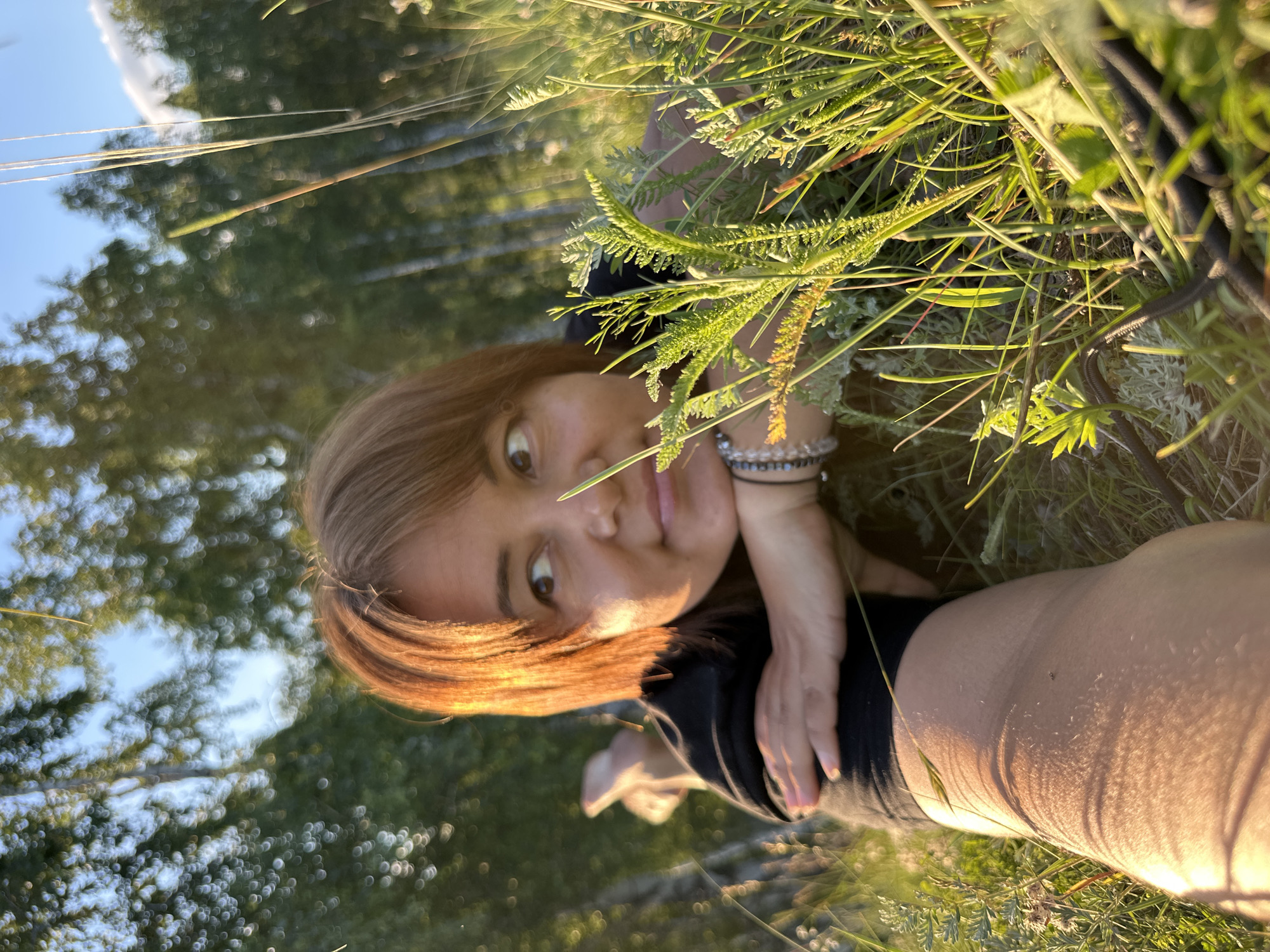 The media file shows a close-up, low-angle shot of a woman lying in tall grass, viewed from above. The woman has light brown hair, is wearing a black shirt, and has a bracelet on her left wrist. Her eyes are wide open, and she appears to be looking directly at the camera. The scene is outdoors, with lush green grass and foliage in the foreground and blurred trees in the background. The lighting suggests it is either early morning or late afternoon, with the sun casting a warm glow on the woman and the grass. The overall mood is casual and serene, with the woman seemingly relaxed in the natural environment. No text is visible in the image.