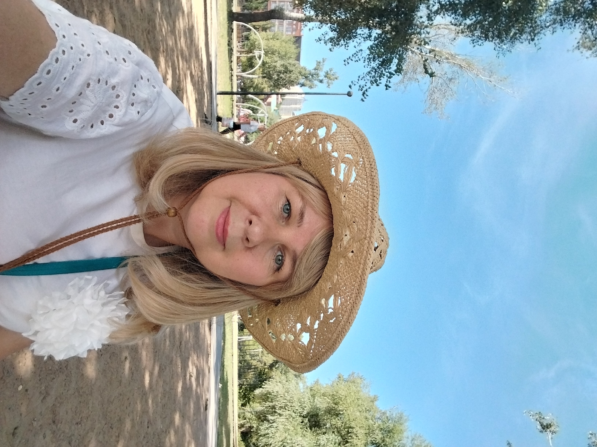 A woman with blonde hair and blue eyes is wearing a straw hat and a white top with eyelet embroidery. She is standing outdoors in a park-like setting with trees and a clear blue sky. In the background, there are buildings and playground equipment. The image appears to be a selfie taken in Orenburg, Russia.