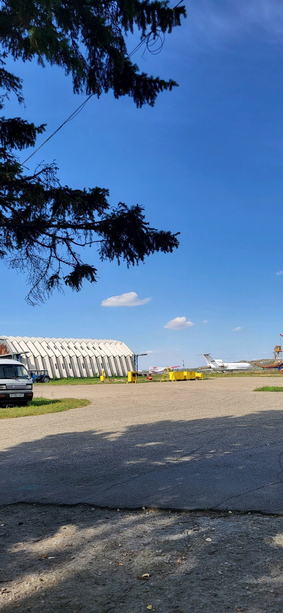 An outdoor, ground-level, vertical, medium shot captures a scene at an airfield in Oskemen, Kazakhstan, under a bright blue sky with scattered white clouds. In the foreground, a dark asphalt surface exhibits shadows from overhead foliage, suggesting it is daytime. A white van, parked on the left side of the frame, faces forward, revealing its license plate, which reads "JF 957 KT." Behind the van and to its left, a blue tractor is partially visible.
Dominating the midground is a large, white, arched hangar with a distinctive patterned facade, stretching horizontally across the image. In front of the hangar, several bright yellow pieces of equipment are present. Further into the background, on the airfield, a white airplane with a distinctive tail is parked, and another smaller red and white aircraft is also visible. The airfield extends into the distance, with a slightly hilly, arid landscape forming the backdrop. The image is devoid of any visible human subjects.